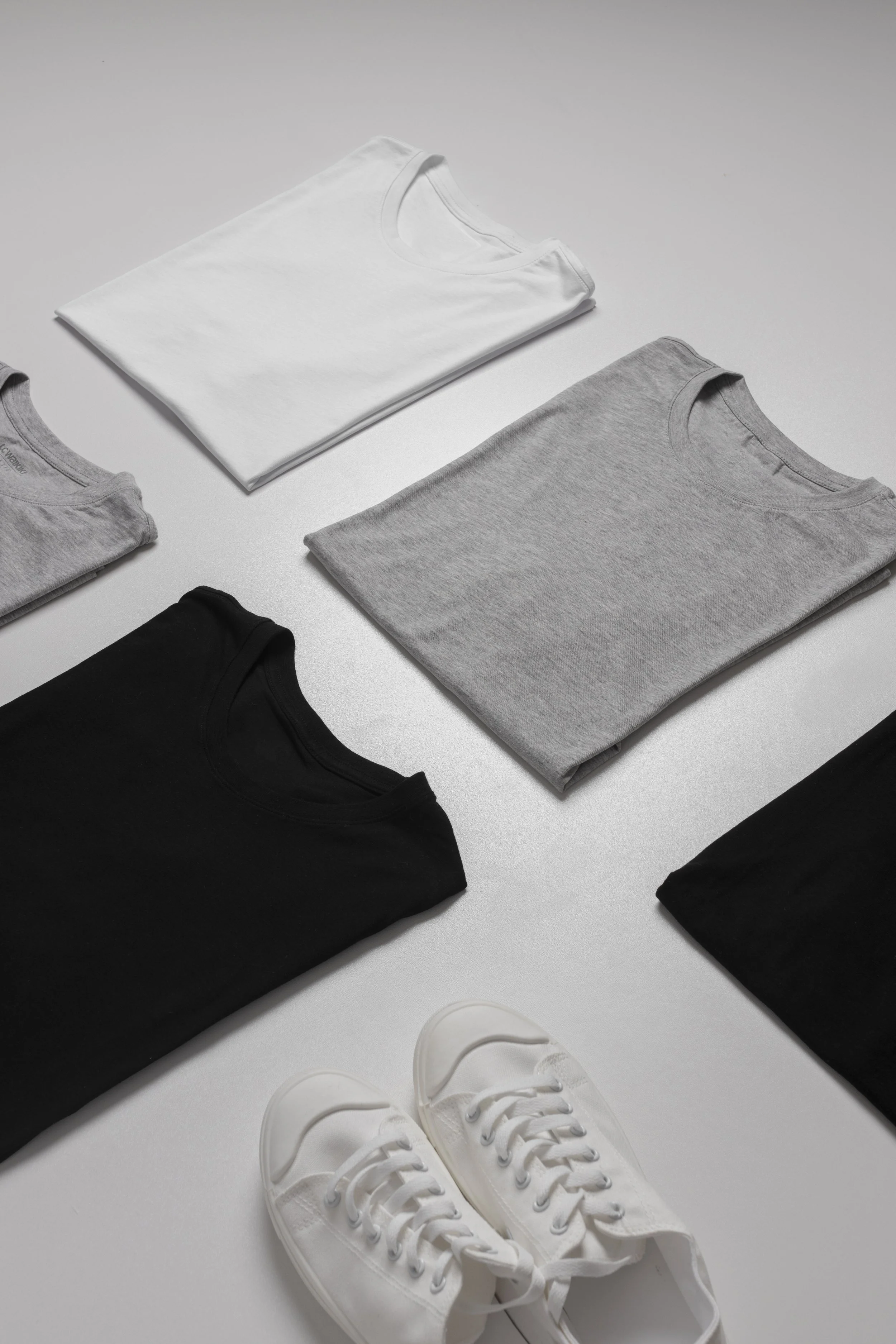 Multiple folded t-shirts in white, gray, black, and light gray colors, along with a pair of white sneakers on a white surface.