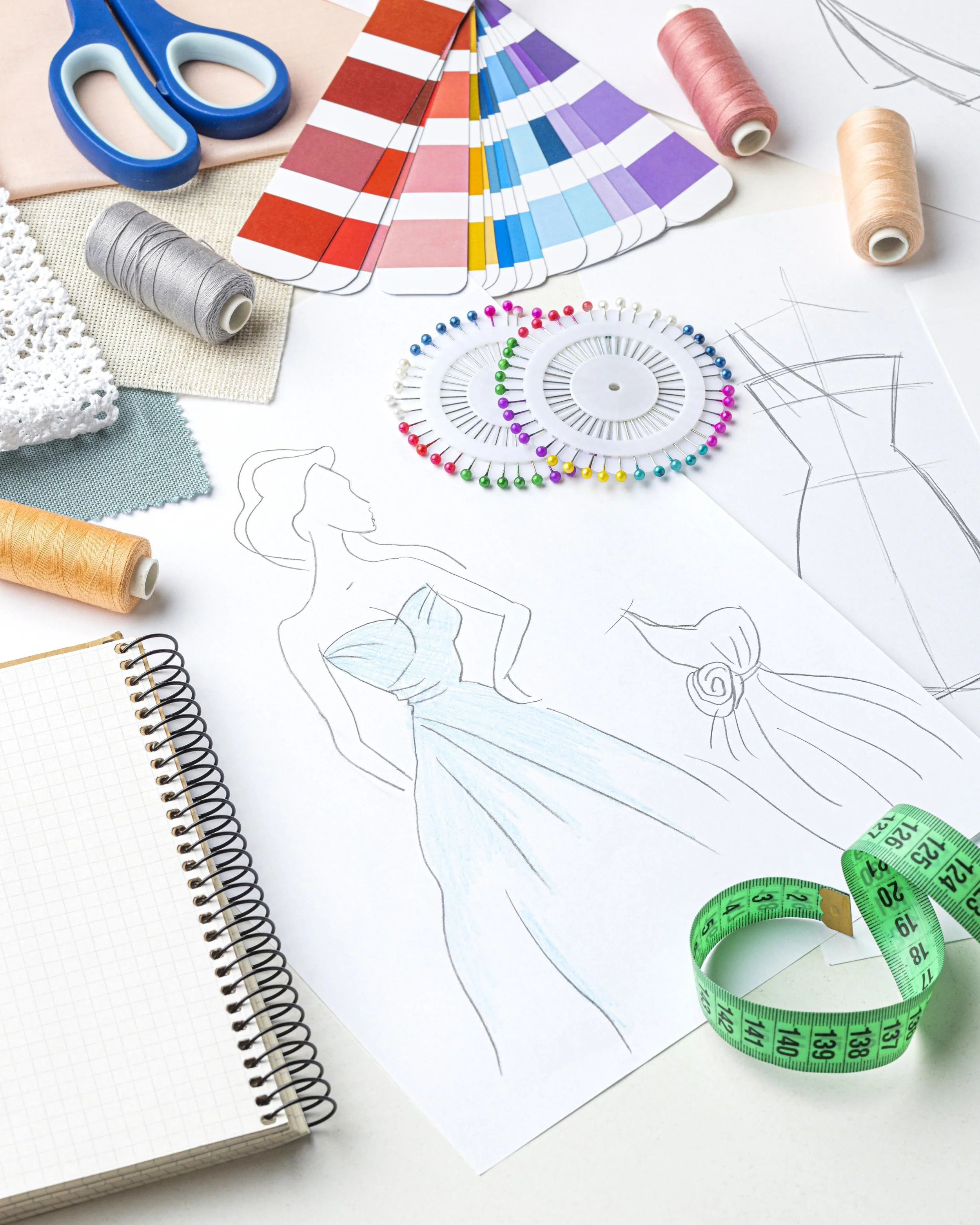 Image of fashion design tools including a color palette fan, sketches of a woman in a dress, spools of thread, sewing scissors, a spiral notebook, fabric swatches, and a measuring tape.