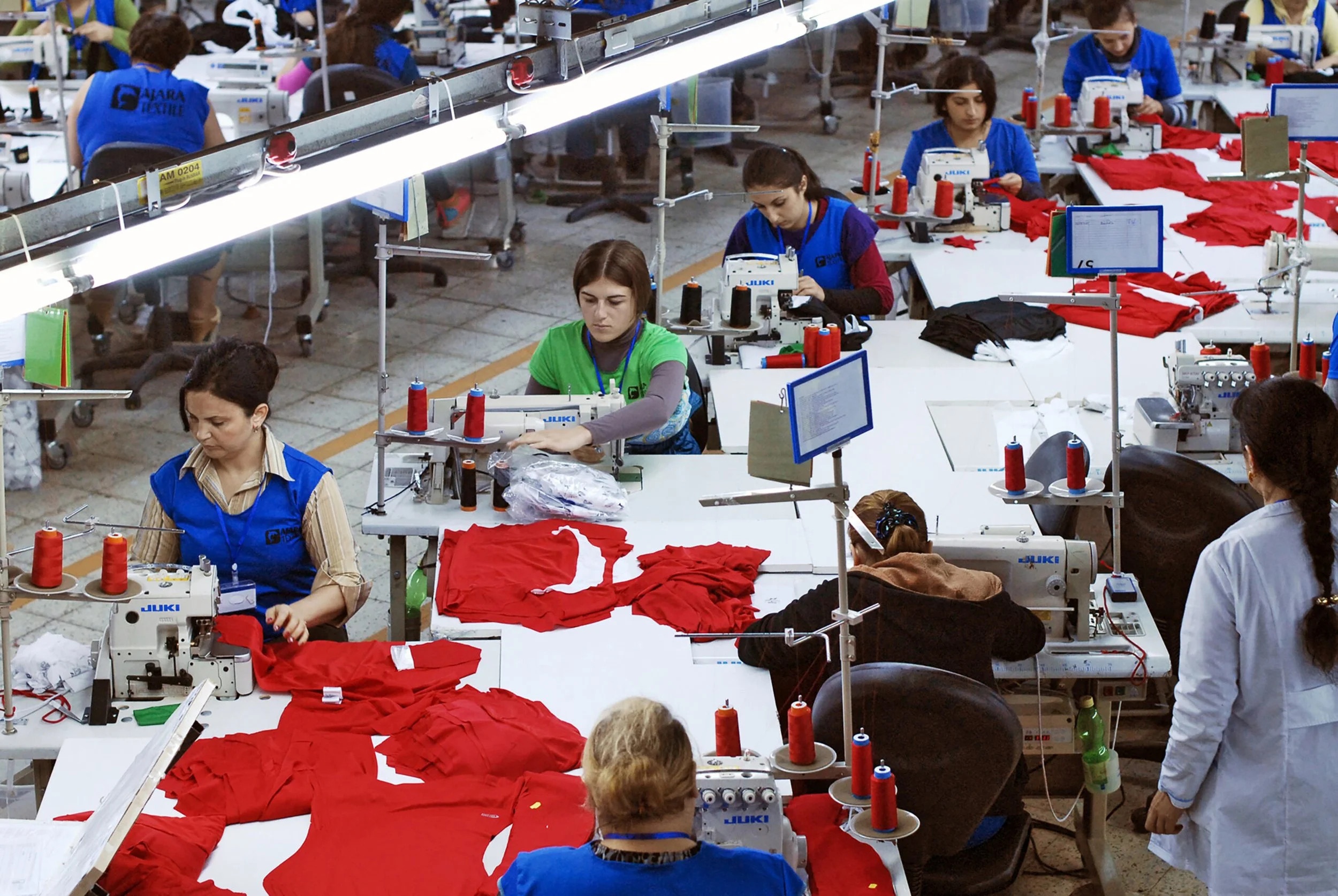 Workers sewing red fabric pieces in a large manufacturing factory.
