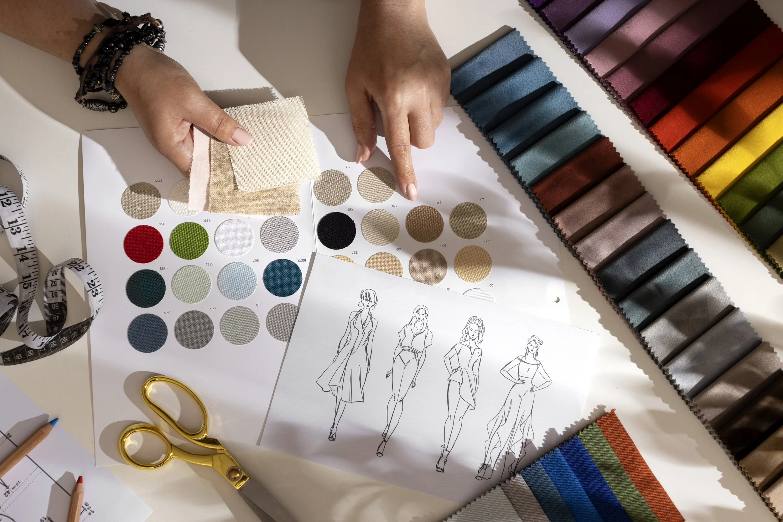 A fashion designer workspace with fabric swatches in various colors, fabric samples, sketches of women in fashion outfits, a measuring tape, and gold scissors.