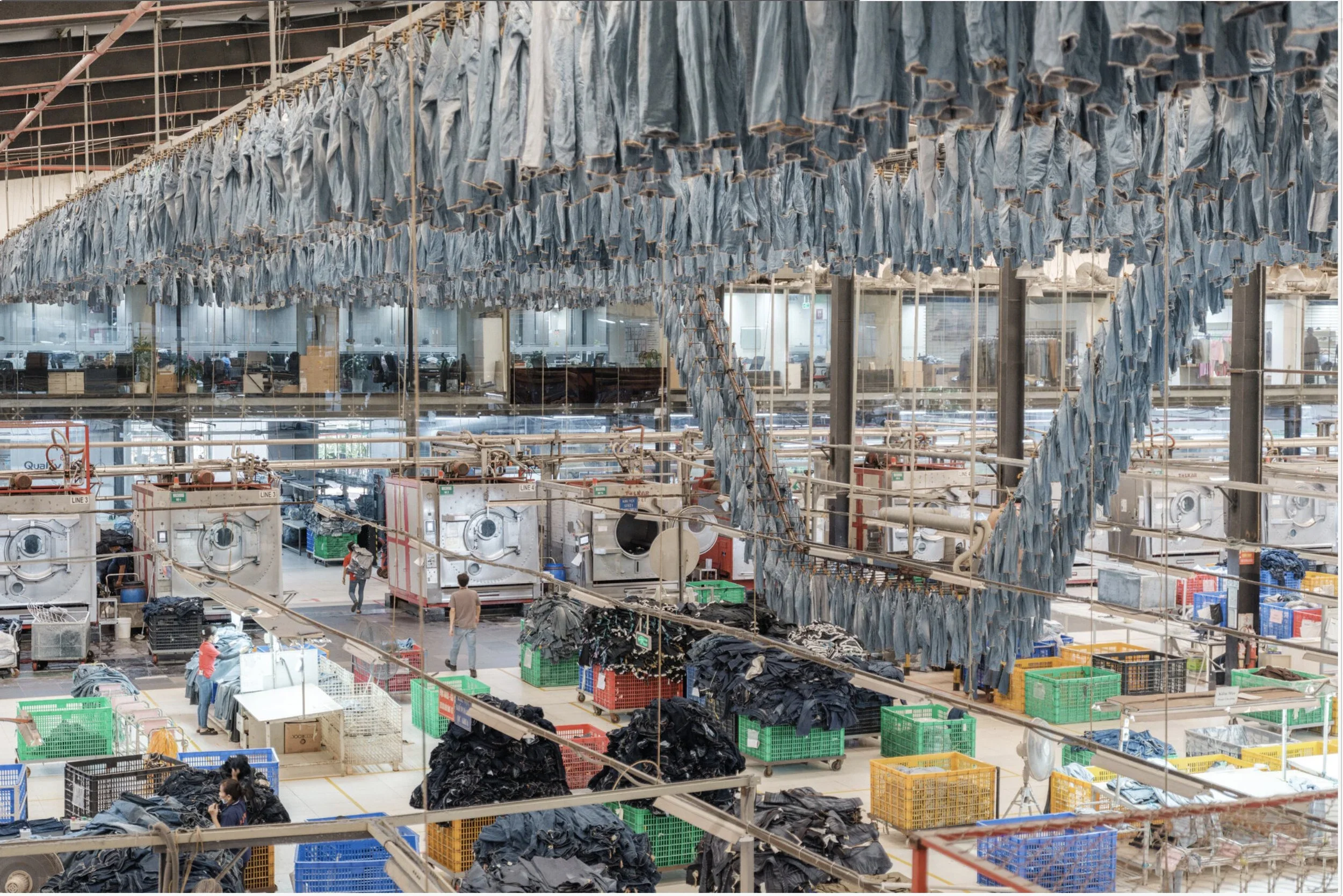 Interior of a large laundromat with clothing hanging from lines overhead and laundry bins on the floor, with some people working.