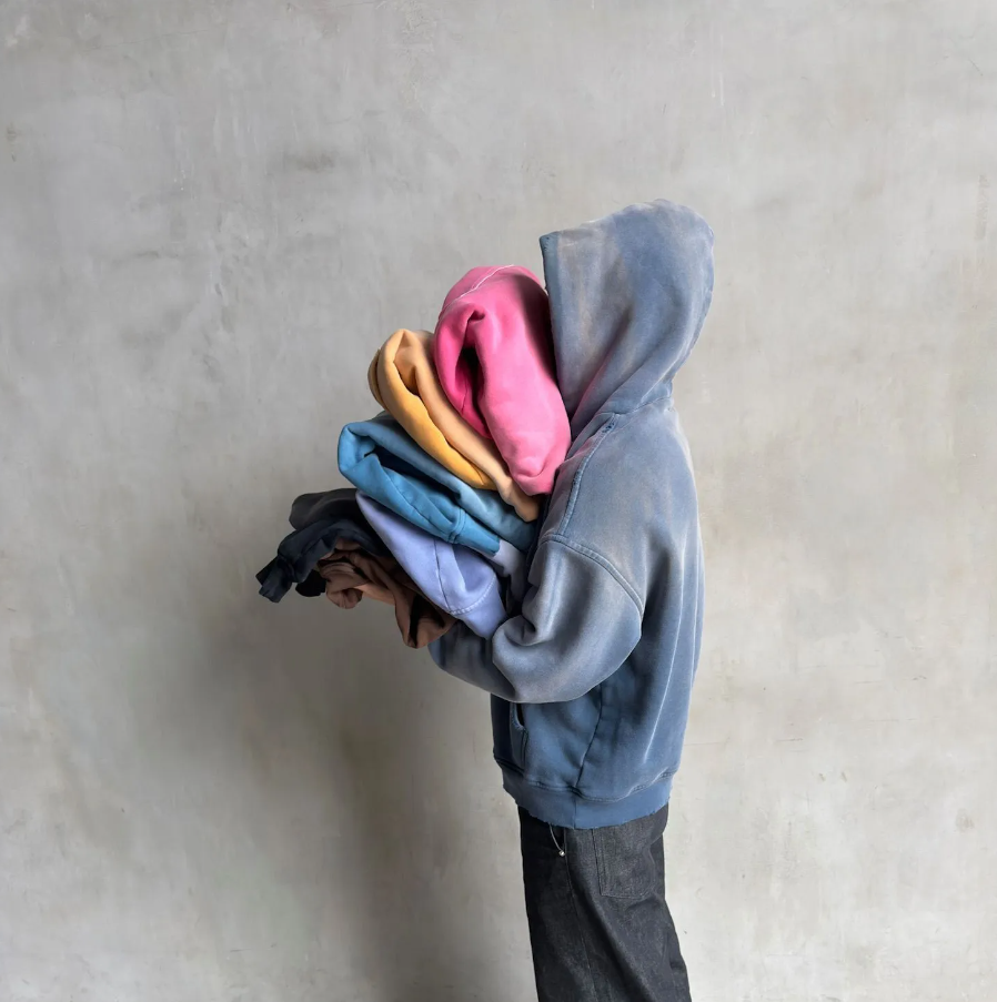 A person wearing a gray hoodie and dark pants is holding a large stack of colorful hoodies or sweatshirts against a plain, light-colored wall.