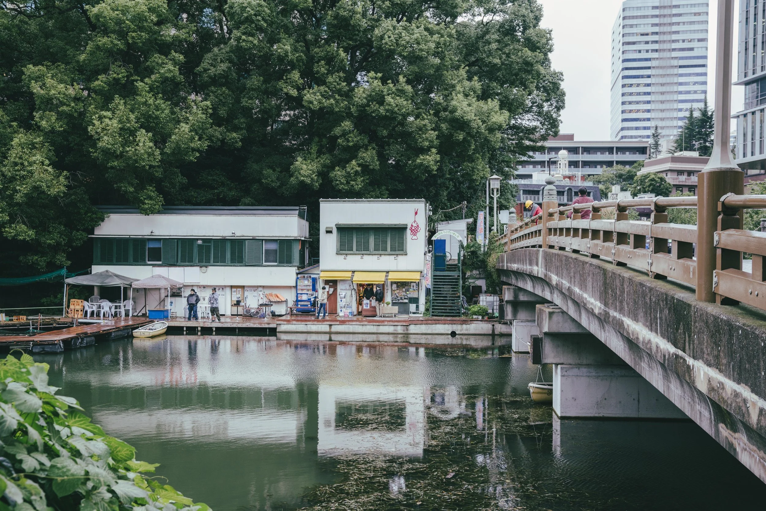 A riverside scene with a bridge and buildings in the background: a small dock with boats, a row of shops or eateries with people nearby, a large green tree, and high-rise buildings in the distance.