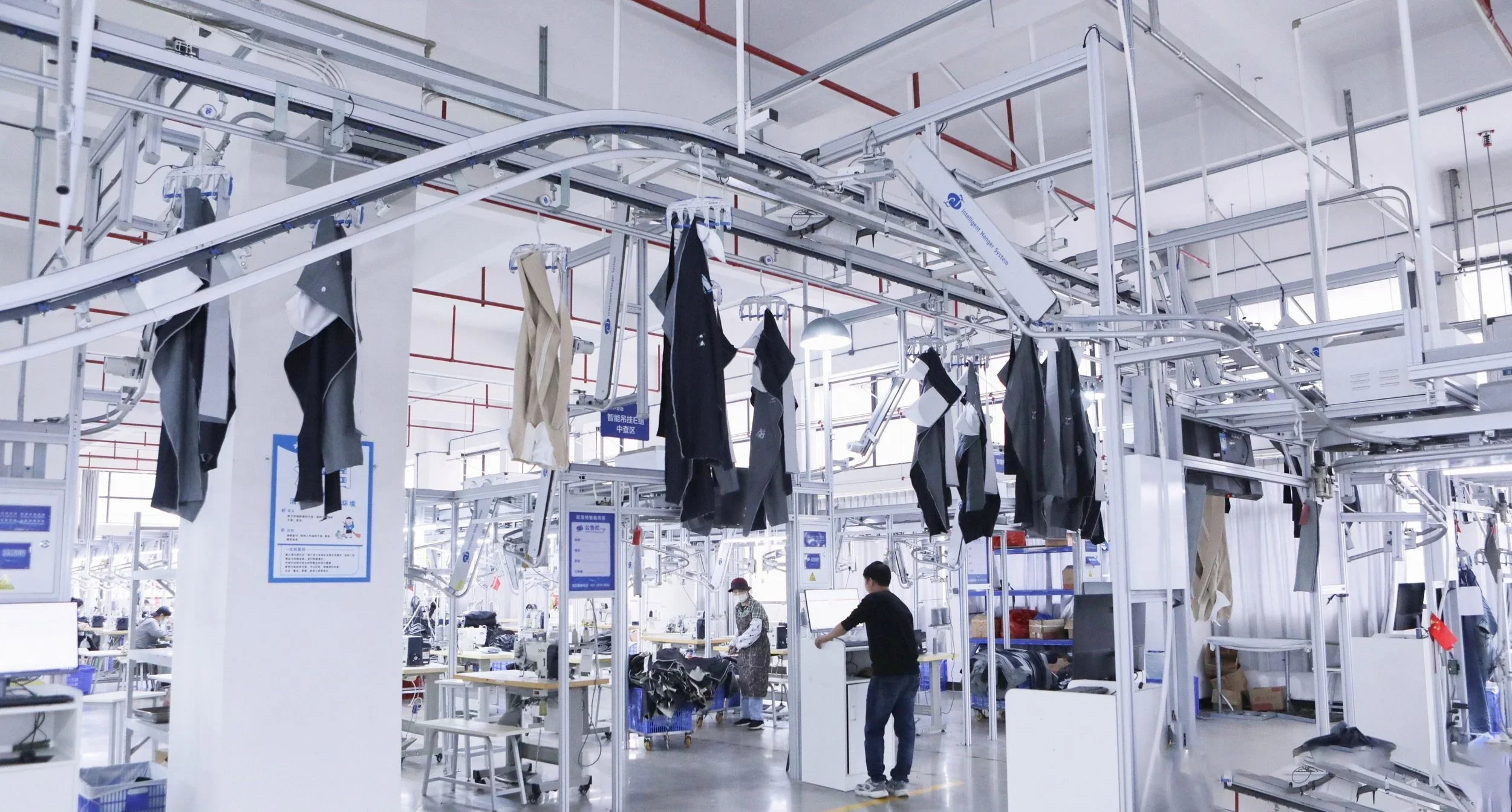 A clothing factory with automated conveyor system hanging clothes, workers operating sewing machines and inspecting garments, with bright lighting and industrial equipment.