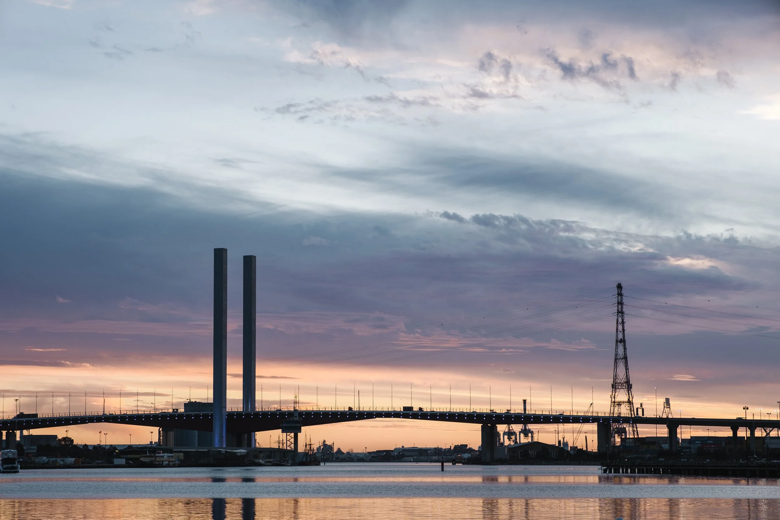 A cityscape during sunset featuring two tall industrial chimneys, a bridge with blue lights, and a transmission tower, with a sky filled with clouds in shades of blue, purple, and pink.