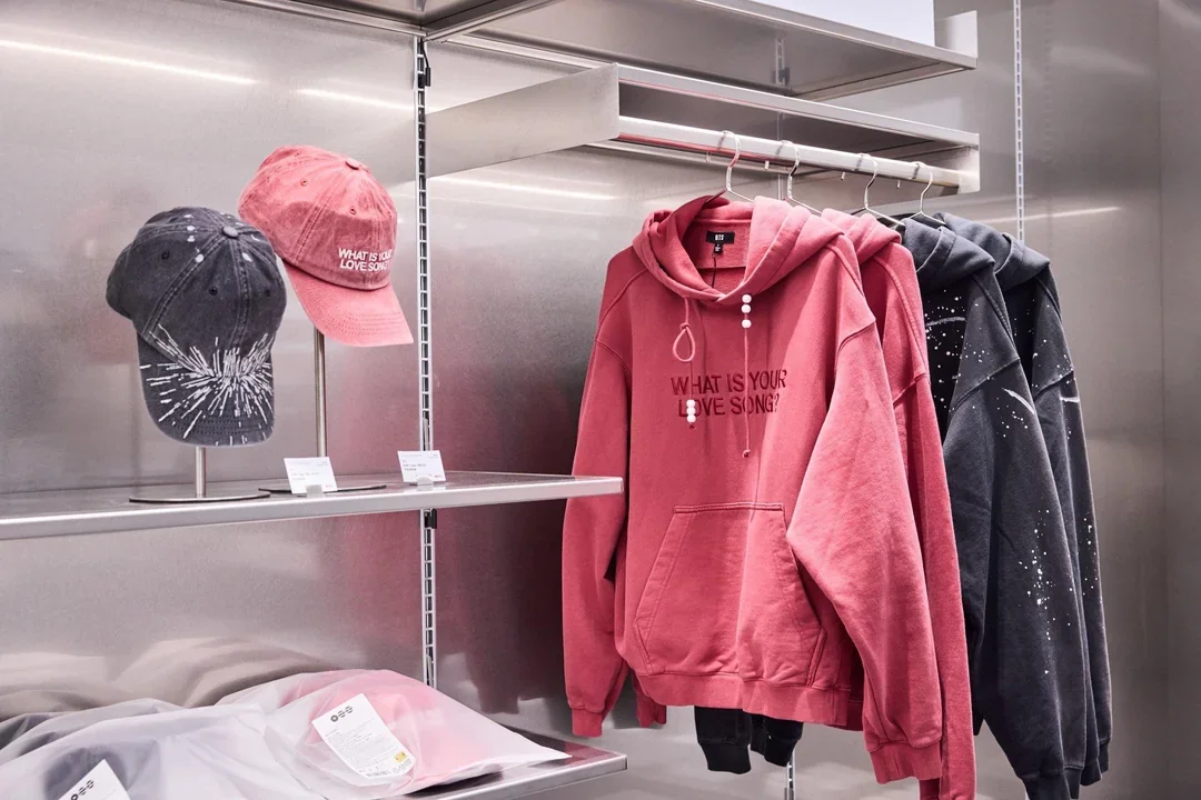 Pink and dark gray hoodies with printed designs on display in a retail store, alongside pink and gray caps on a rack.