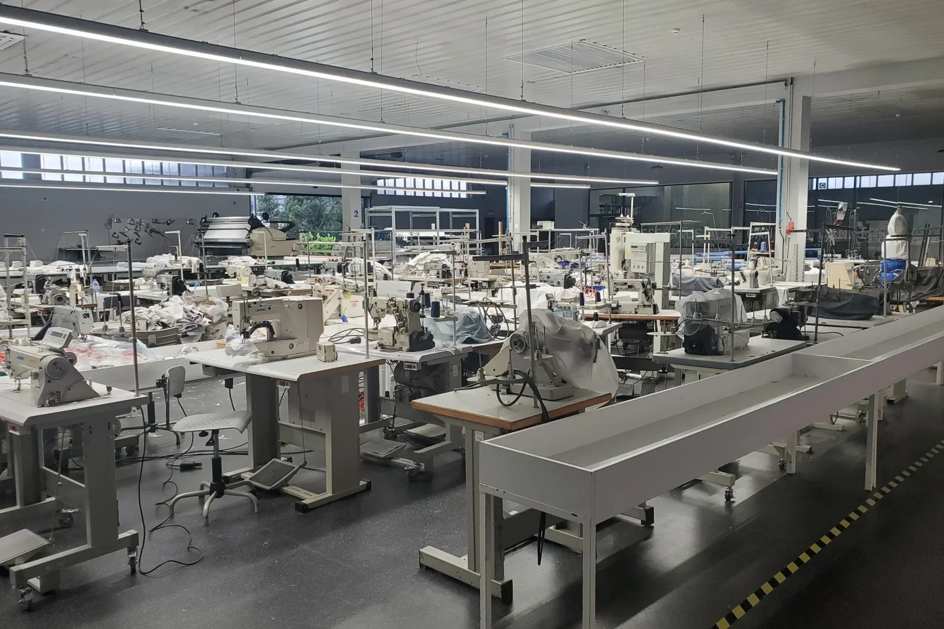 Empty factory or workshop with rows of sewing machines and worktables, illuminated by long overhead lights, with some fabric and materials scattered on the tables.