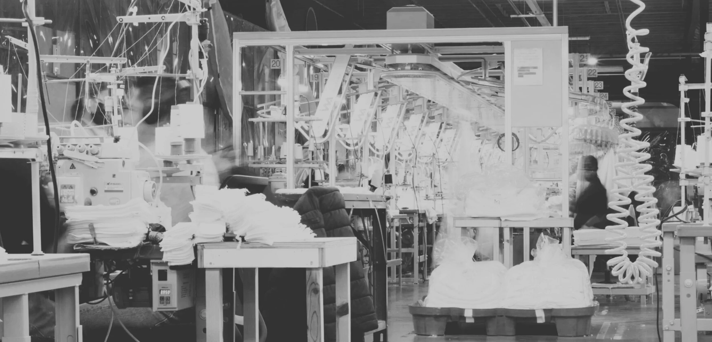 This is an image of a crowded manufacturing or industrial workspace with equipment, pipes, and machinery. There are tables with fabric or paper materials, and various tools and supplies scattered around.