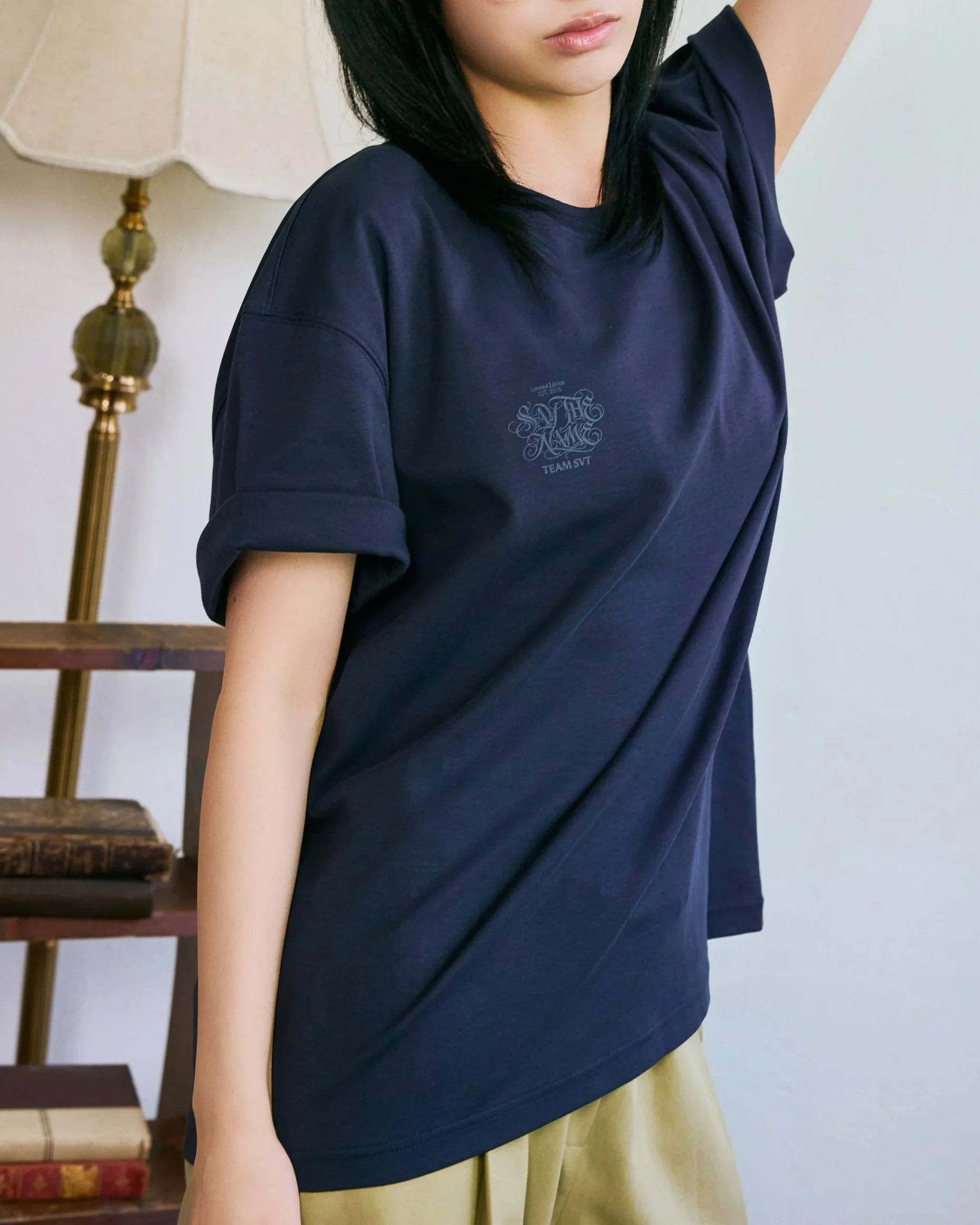 A woman wearing a navy blue T-shirt with a small logo on the chest, standing indoors next to a brass floor lamp with a beige lampshade and wooden stairs.