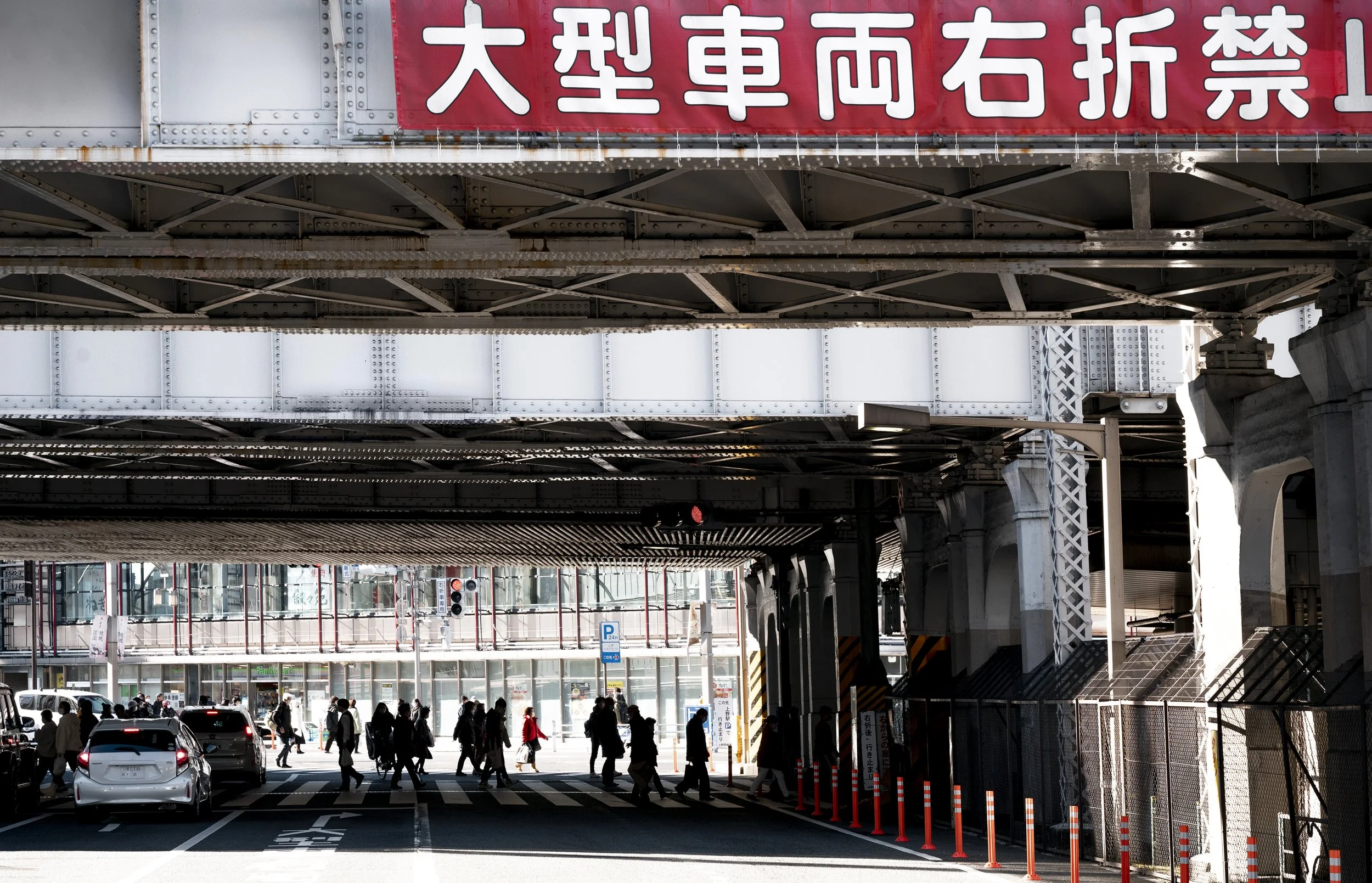People walking beneath a bridge with a red Japanese sign, cars on the street, and a parking sign in an urban area.