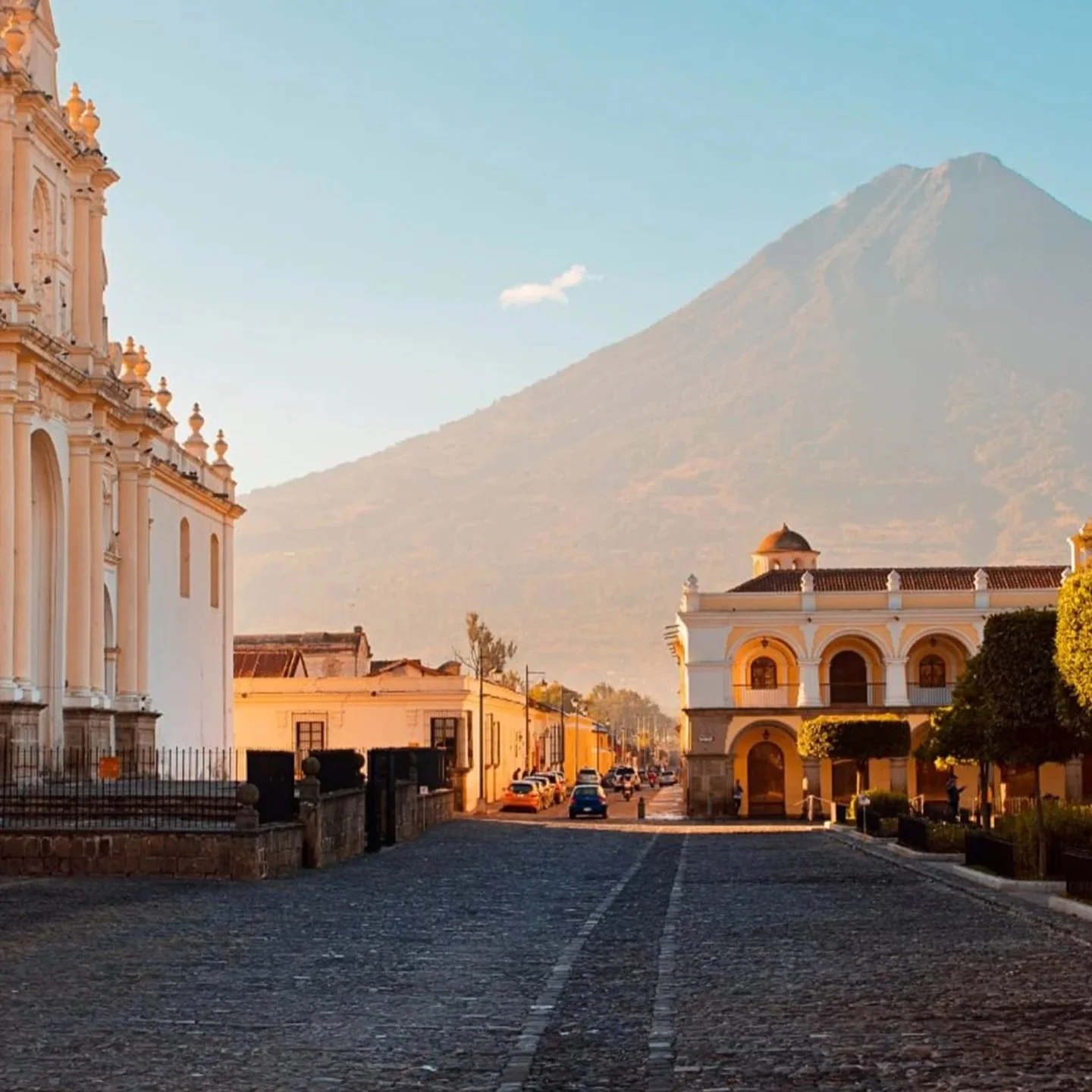A cobblestone street in a historic town with colonial-style buildings, a church on the left, and a white building with arches on the right, with a large volcanic mountain in the background, during sunset.