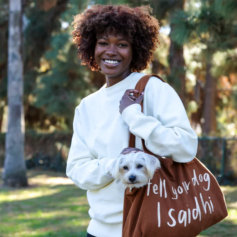 A young woman smiling outdoors, holding a small dog in a bag and carrying a brown tote bag with the text "tell your dog I said hi." She has curly hair, wears a white sweatshirt, and stands in a park with trees.