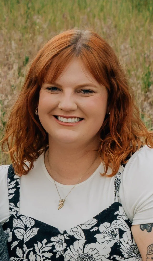 Portrait of a smiling woman with red curly hair, wearing a white shirt and a black floral dress, outdoors in a grassy area.
