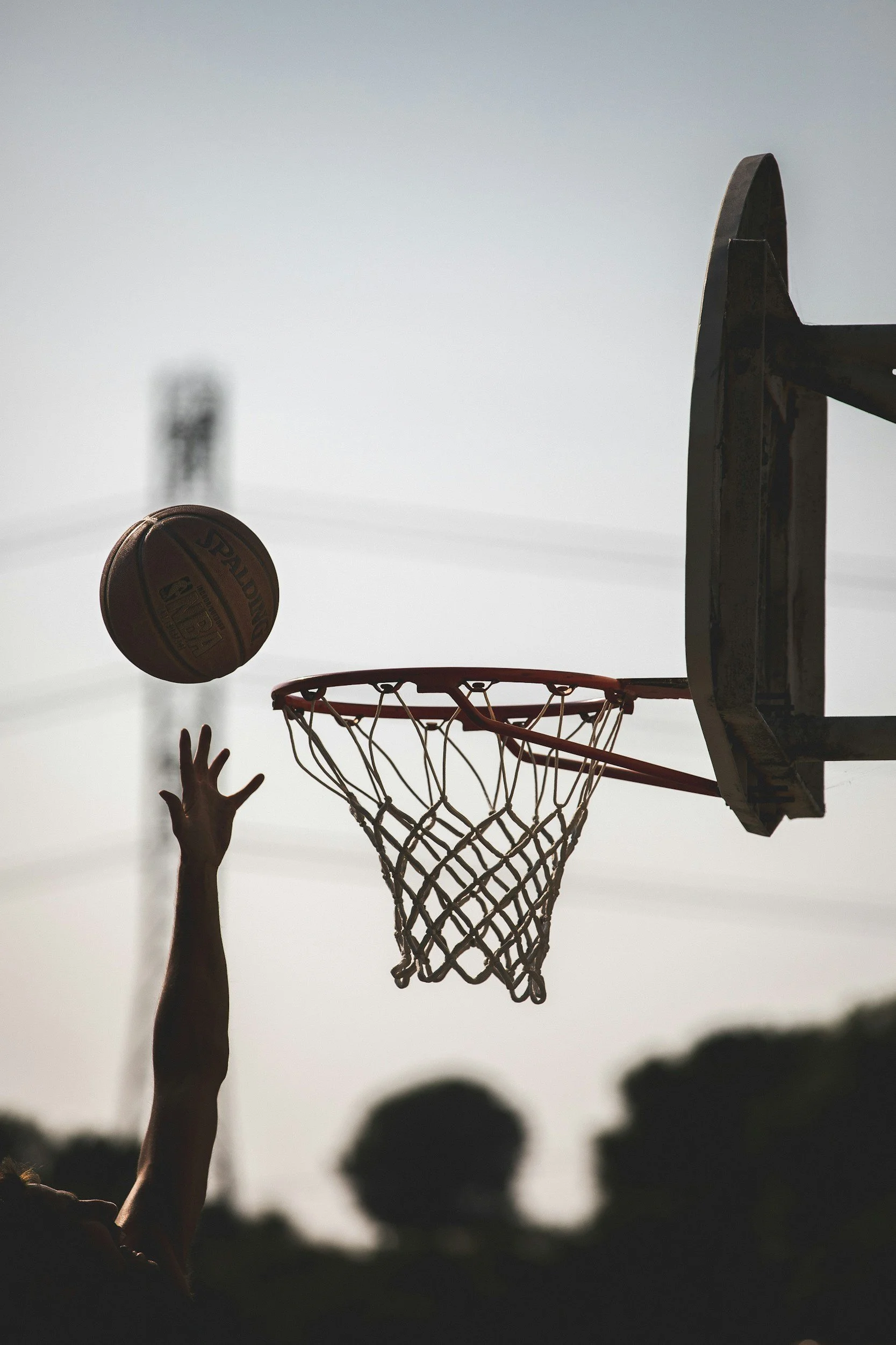 Children playing basketball outdoors during sunset, shot from below the hoop showing hands and a basketball in mid-air, with silhouettes of trees and the sky in the background.