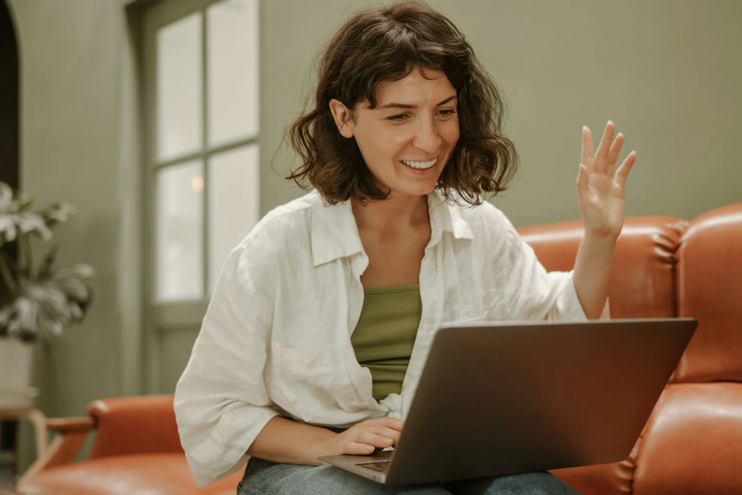 A woman with short, curly brown hair smiling and waving while sitting at a table with a laptop in front of her, indoors near a window.
