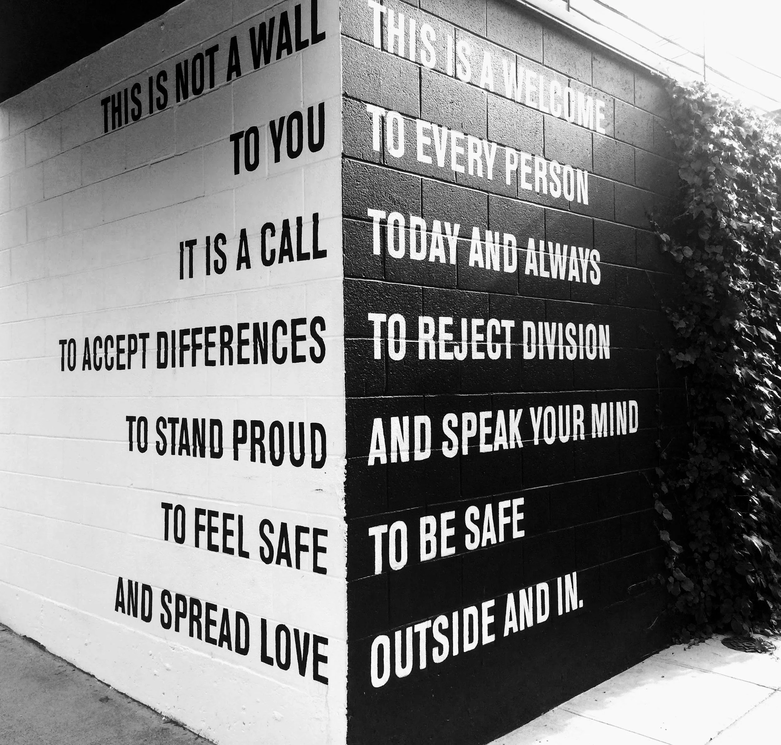 Black and white photo of a corner of a brick wall with multiple words and phrases written on it with spray paint, promoting love, acceptance, and understanding, including phrases like "This is not a wall," "to you," "love," and "speak your mind."