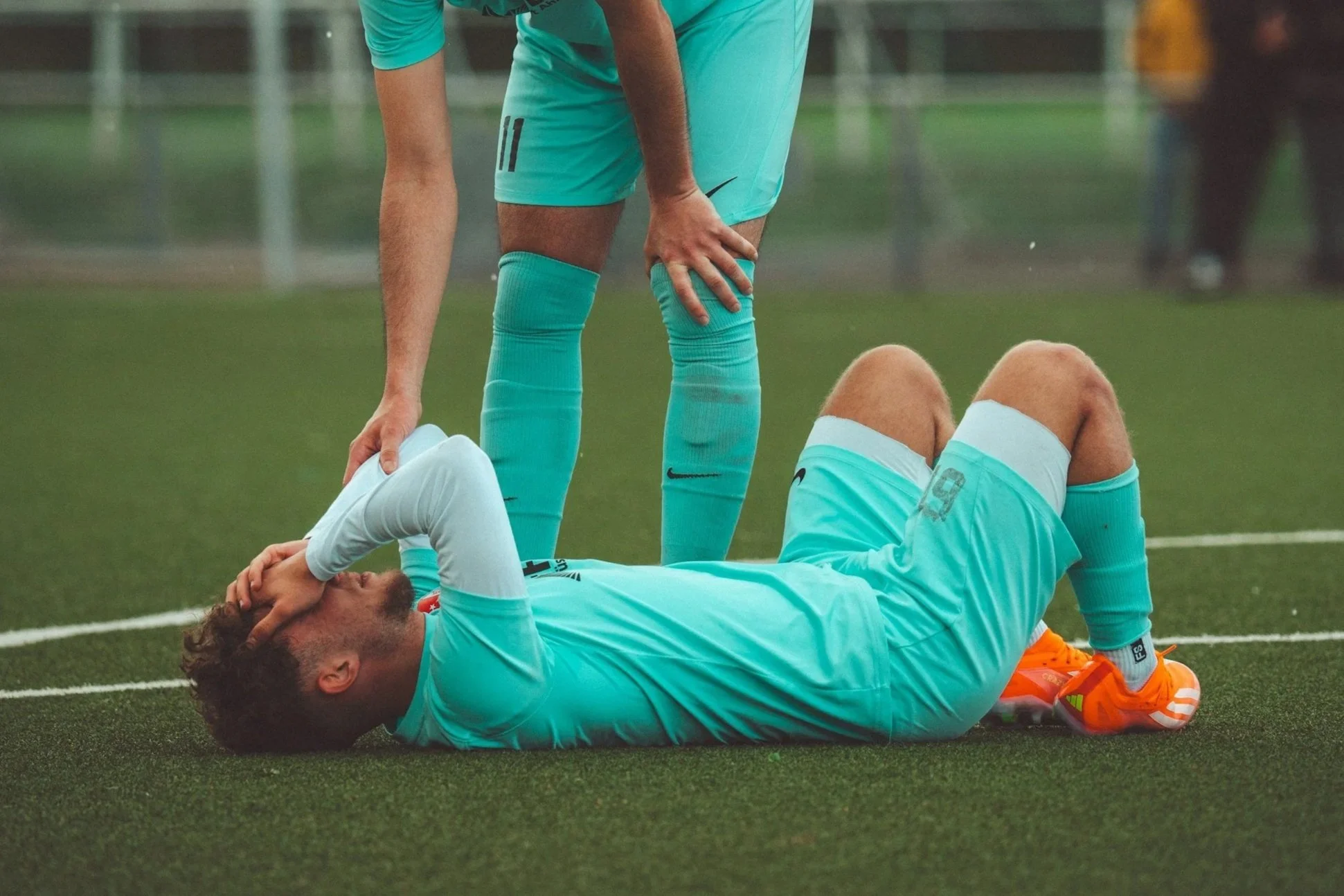 A soccer player lying on the field with a hand on his forehead and another on his face, while a teammate stands over him in a moment of concern during a game.