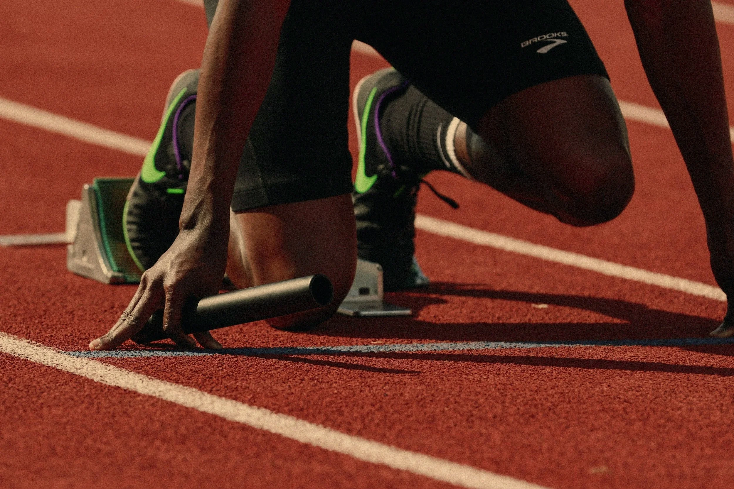 A male athlete in a starting position on a red running track, ready to race, with racing shoes and a starting block.