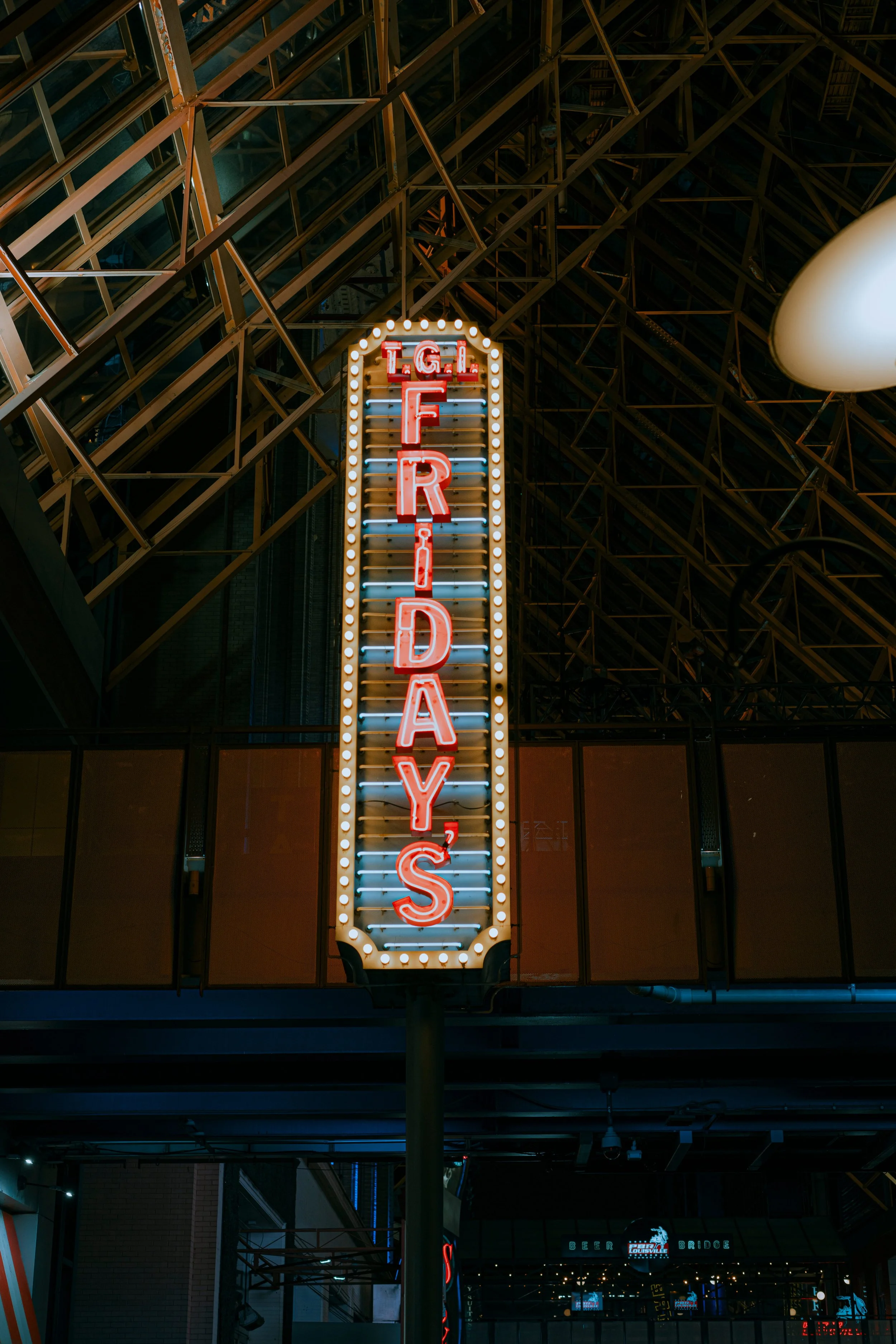 A neon sign that says 'Texas' vertically, with the letters lit up in red, surrounded by a border of small round light bulbs, hanging outside a building with a metal framework and glass windows at night.