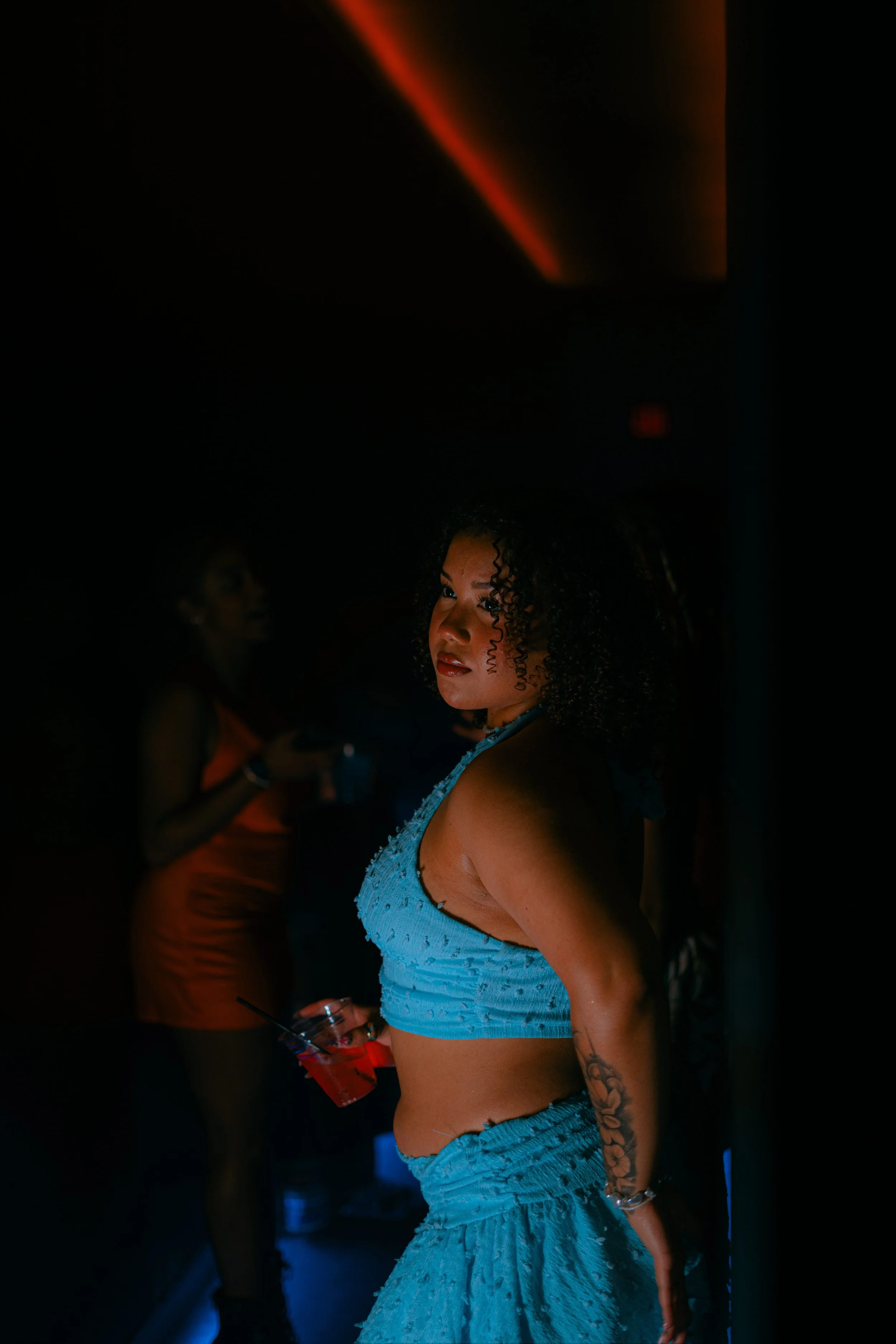 A woman with curly hair wearing a blue sleeveless dress stands in a dark room with red lighting, holding a drink and looking to the side. Two women in orange dresses are blurred in the background.