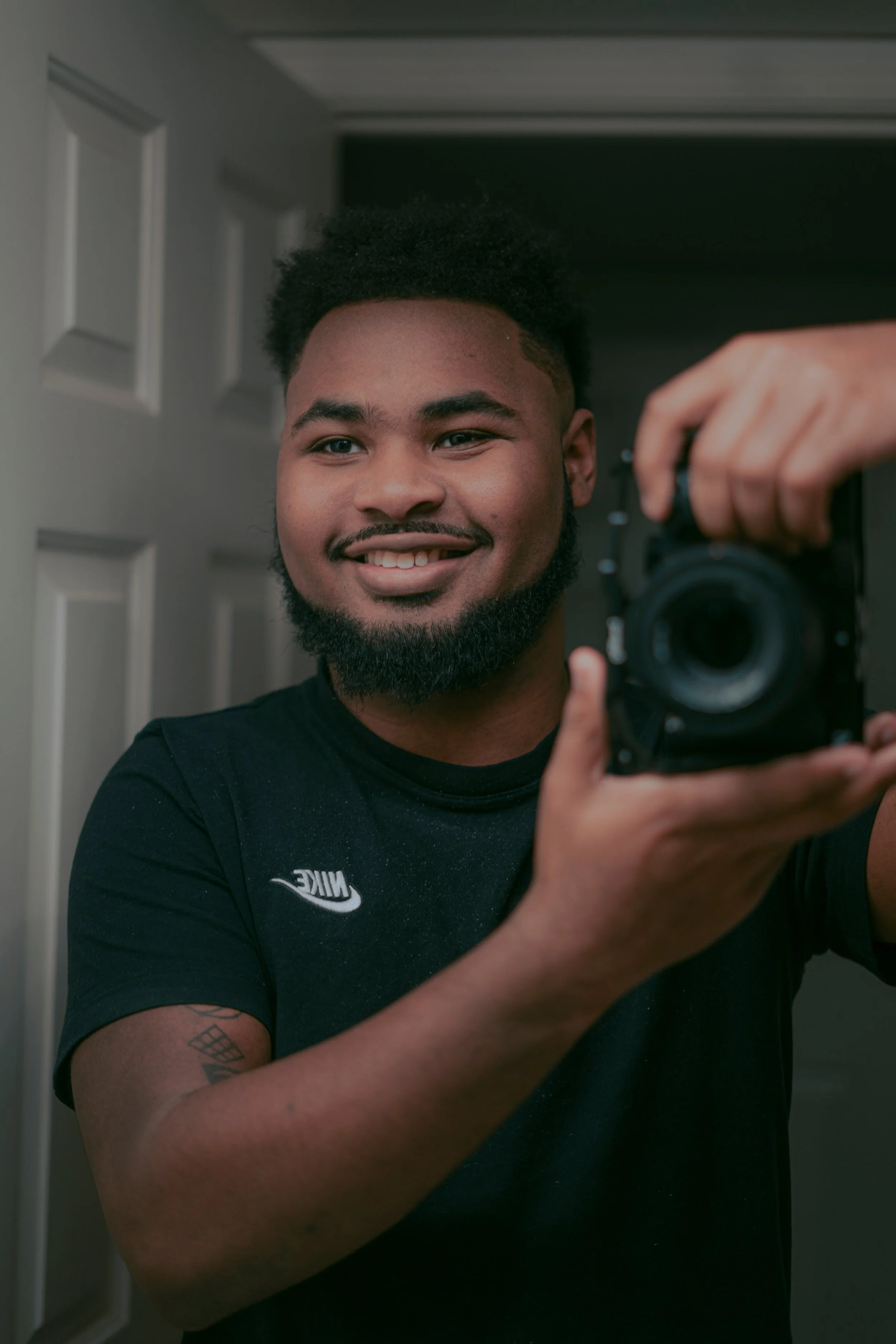 A young man with a beard and short curly hair taking a selfie in front of a mirror, smiling, holding a camera. He is wearing a Nike black t-shirt with tattoos on his left arm.