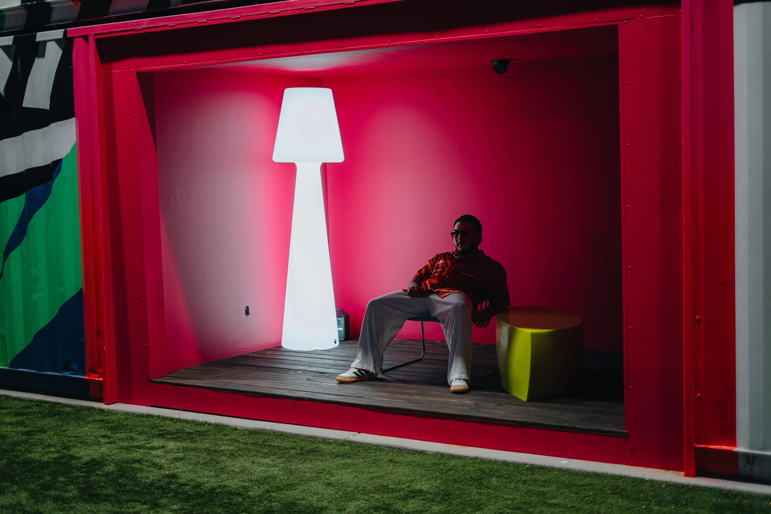 A person sitting in a pink booth with a large white lamp behind them, colorful walls, and seating area outside on the grass.
