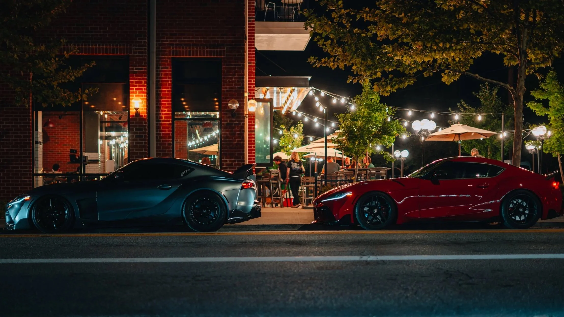 Night scene of a restaurant with outdoor seating illuminated by string lights, with two sports cars parked in front, one black and the other red, and people dining inside.