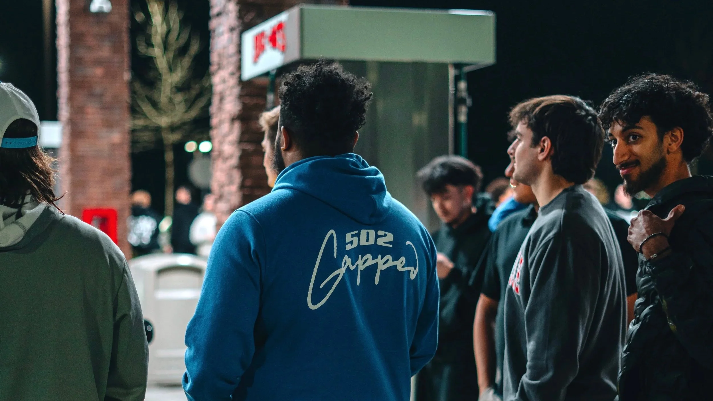 A group of young men standing together outdoors at night, some with curly hair and wearing hoodies, engaged in conversation.