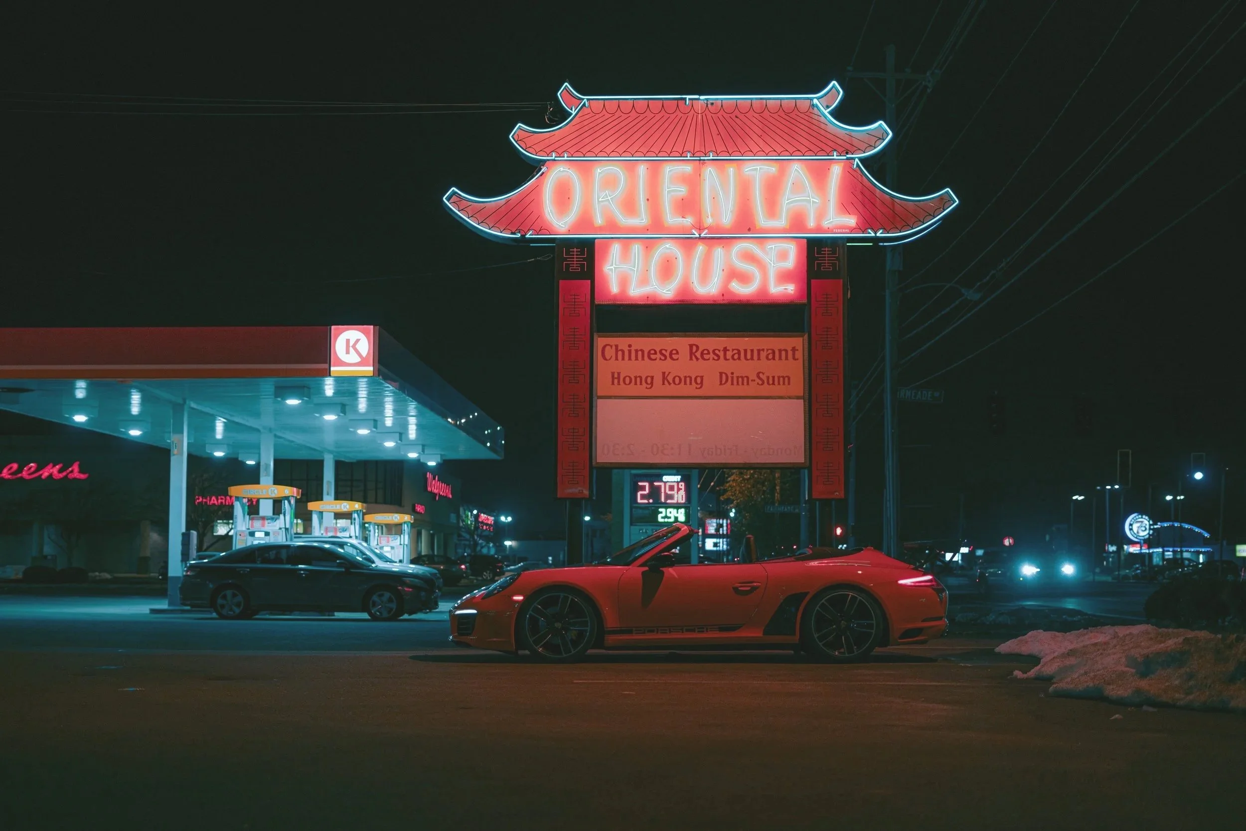 Night shot of a neon sign for Oriental House, a Chinese restaurant, featuring a pagoda-style roof outline and red lettering, with a gas station and sports cars in the foreground.