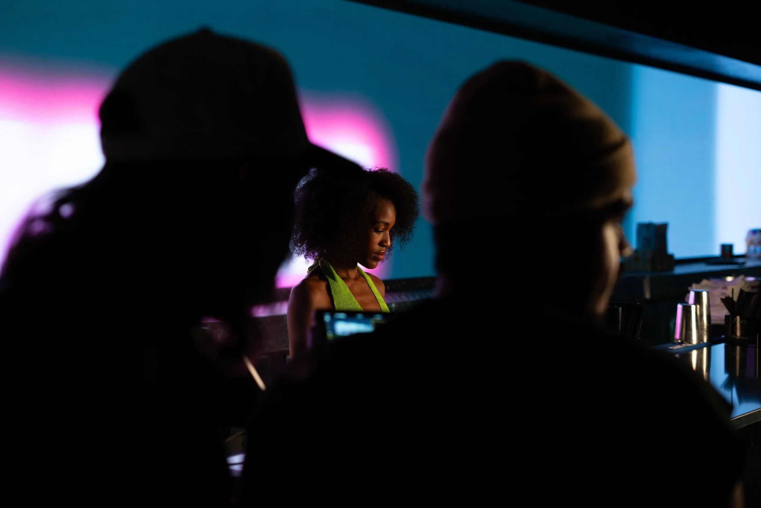 Silhouettes of multiple people in a dark room with a woman in the background illuminated by colored lights, possibly at a nightclub or bar.