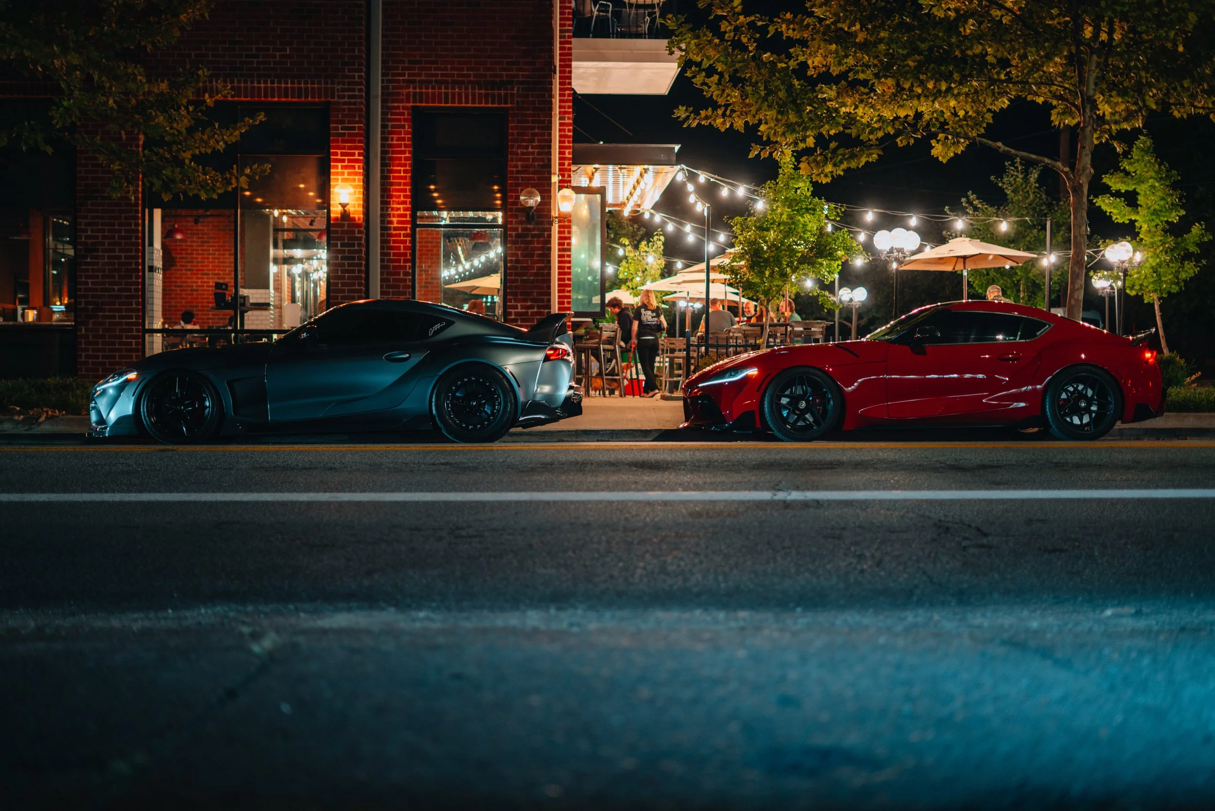 Night view of two sports cars parked on the street in front of a restaurant with outdoor seating, string lights, and trees with green leaves.