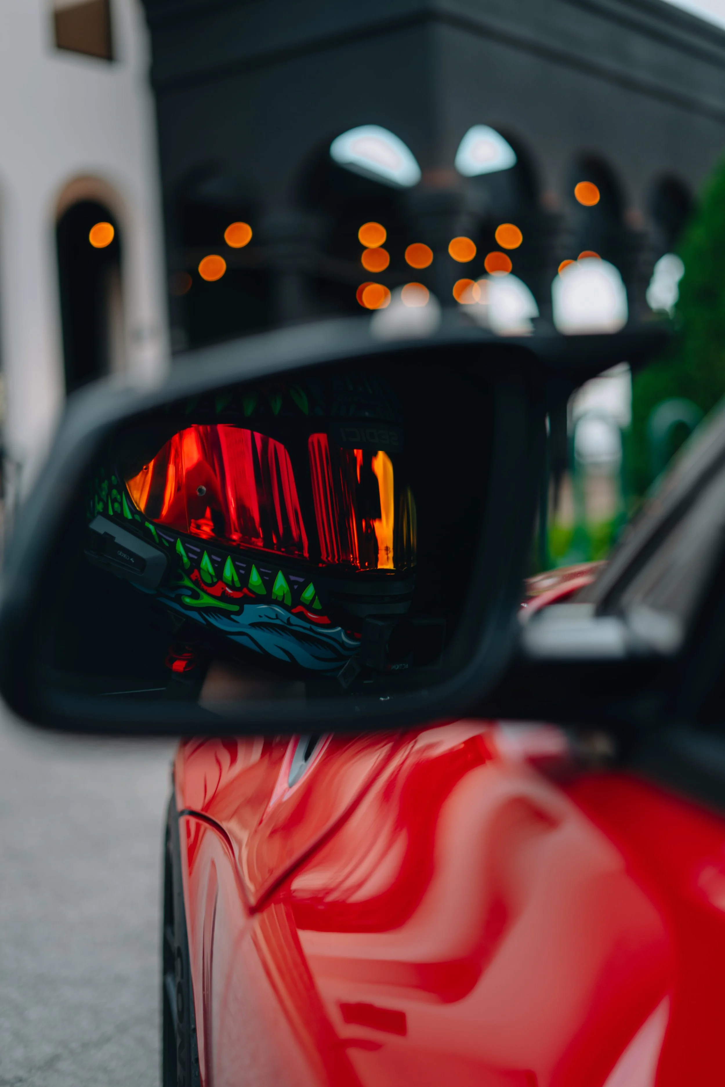 A close-up view of a red sports car seen through its side mirror, reflecting a colorful helmet with a dinosaur design and tinted visor.