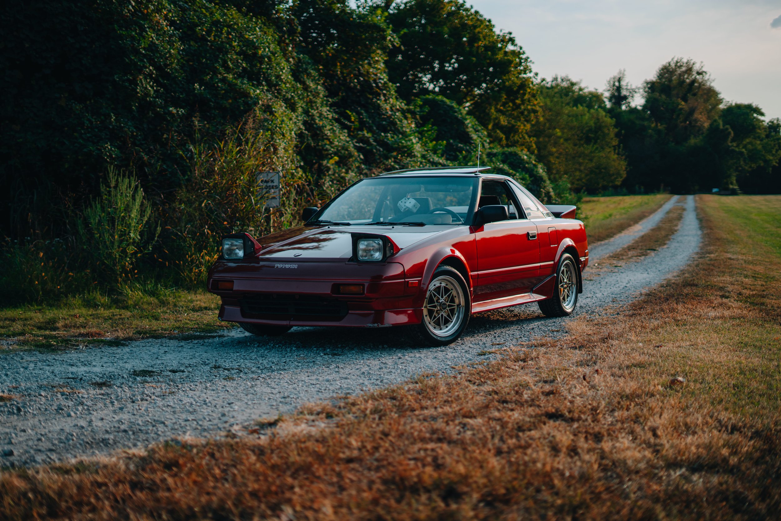 Red vintage Toyota sports car parked on a gravel path beside lush green trees and shrubbery during daytime with cloudy sky.