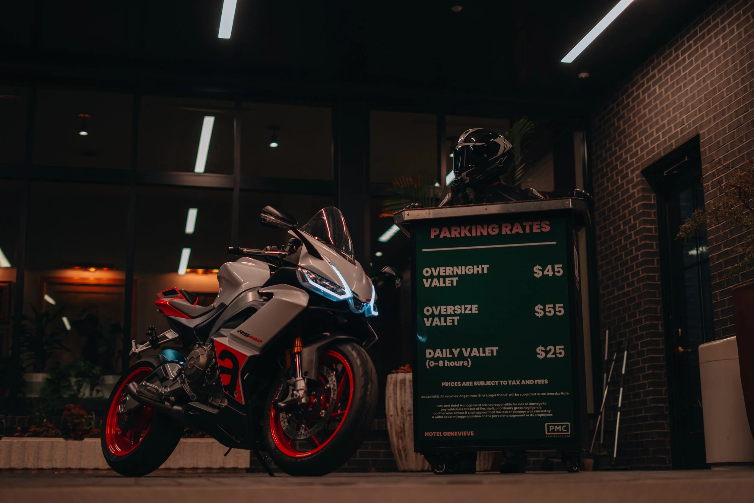 A white motorcycle with red accents parked outside a building at night. In the background, there's a green parking rates sign with prices for overnight, valet, and daily parking. The building has large glass windows and a brick wall.