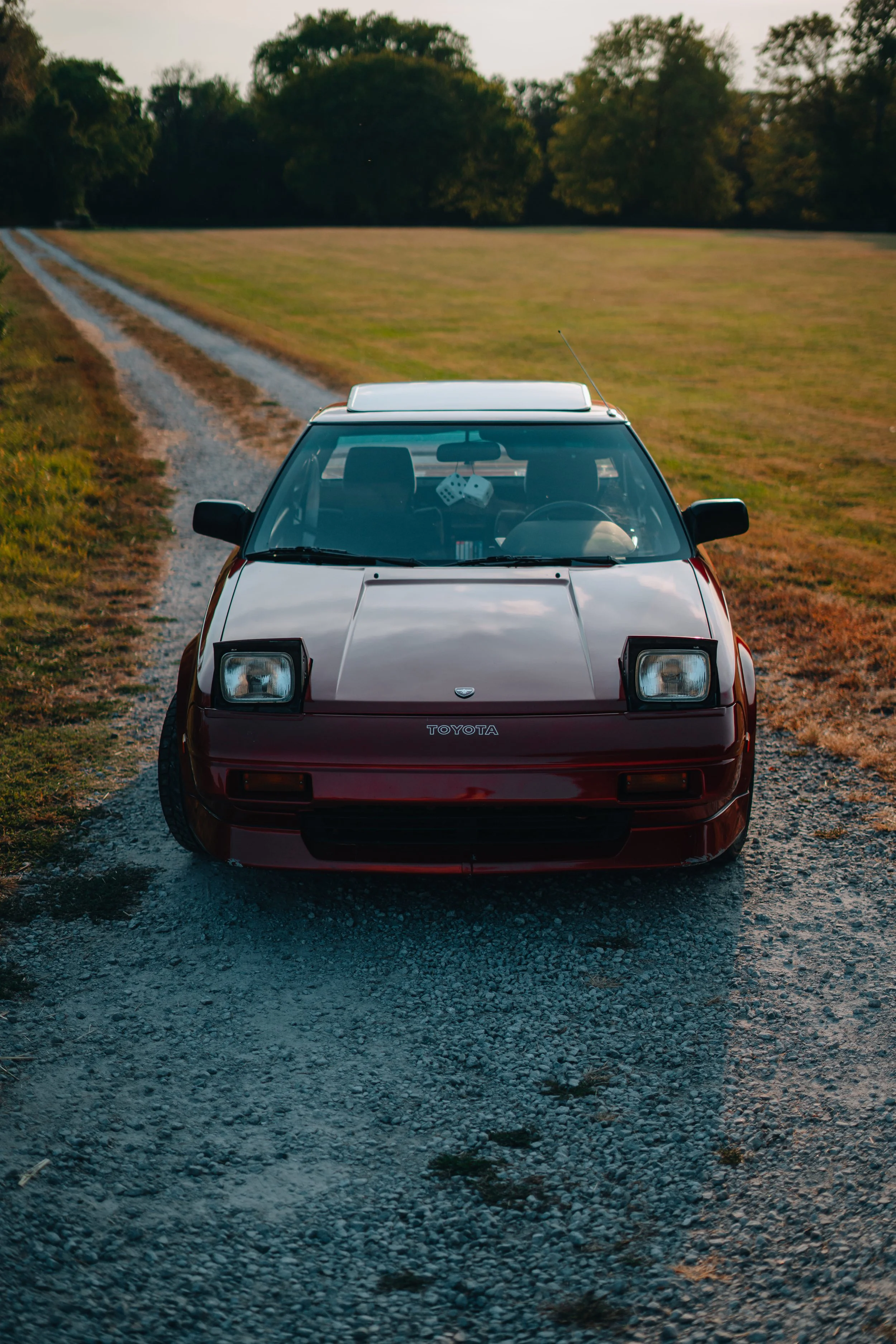A red Toyota car with a white roof is on a gravel road surrounded by green fields and trees in the background.