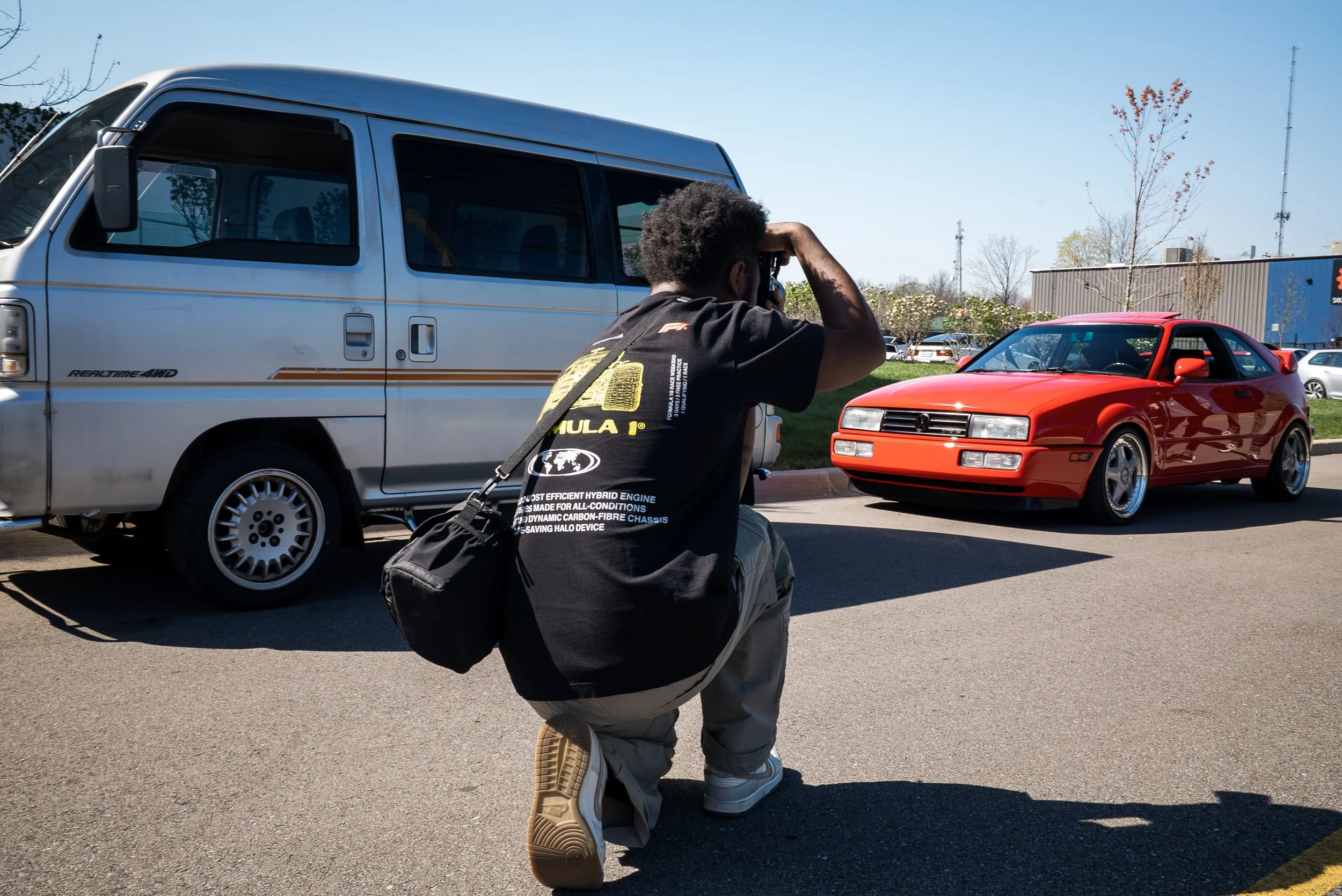 A person is kneeling and taking a photograph of a red sports car parked next to a white van in an outdoor parking lot.