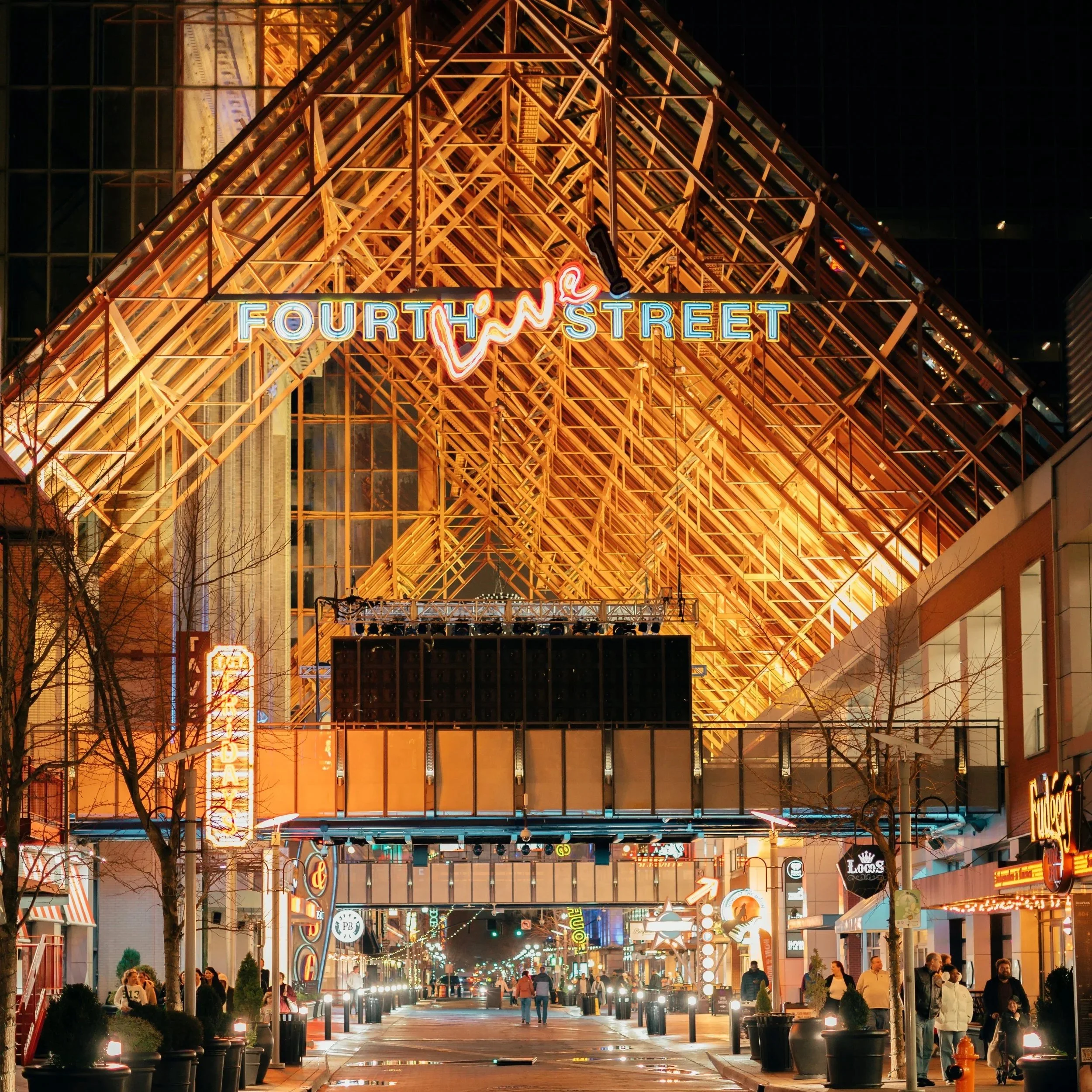 Night view of Fourth Street with a large wooden A-frame structure, colorful neon signs, and people walking along a lively city street.