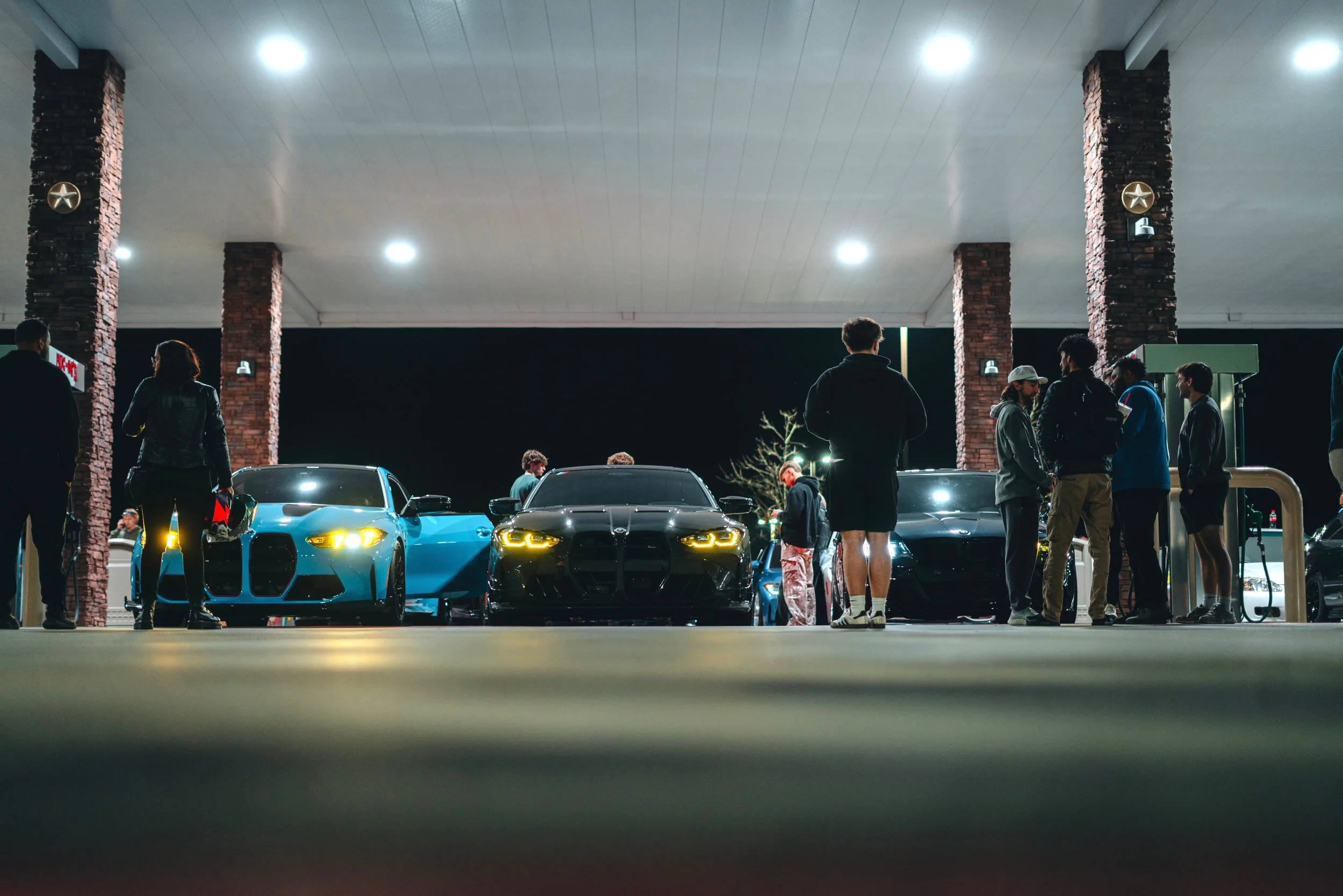 A group of people gathered around three sports cars at a car meet at night, with brick pillars and bright overhead lights in the background.