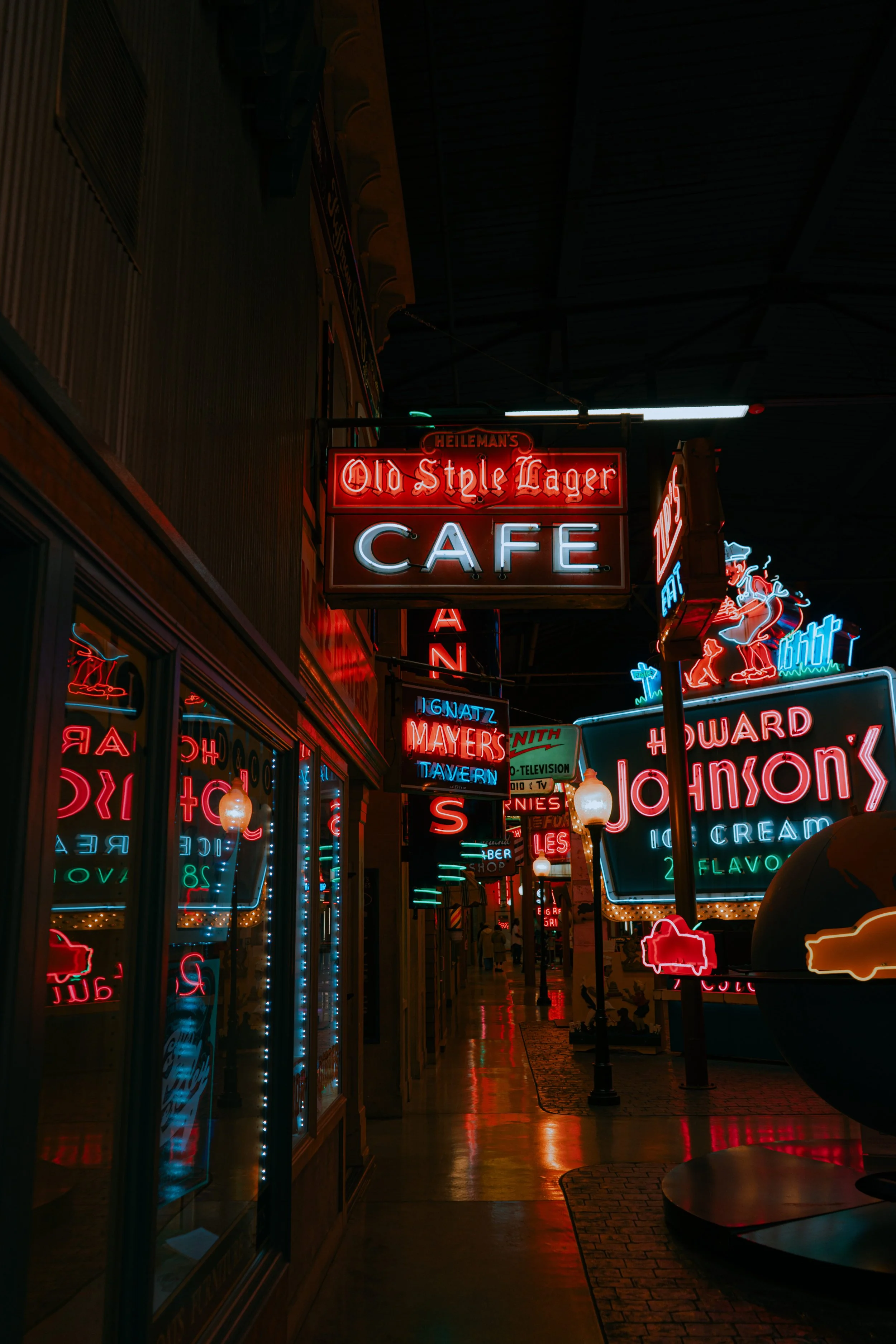 Nighttime view of a neon-lit street with signs for a cafe, tavern, and ice cream shop, featuring vibrant red, blue, and orange neon lights.