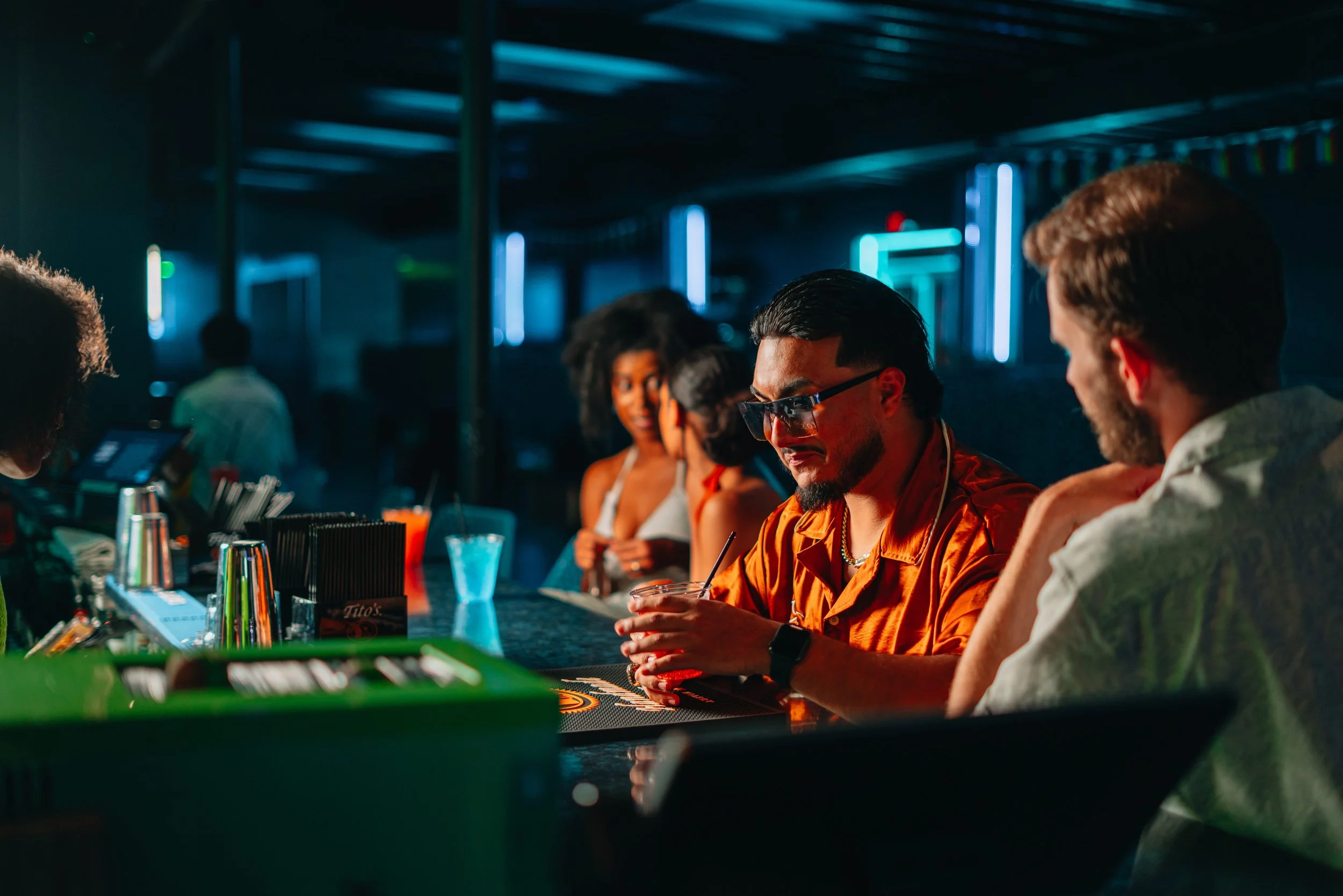 Group of people sitting at a bar counter in a dimly lit room, some engaging in conversation, others enjoying drinks, with neon lights visible in the background.