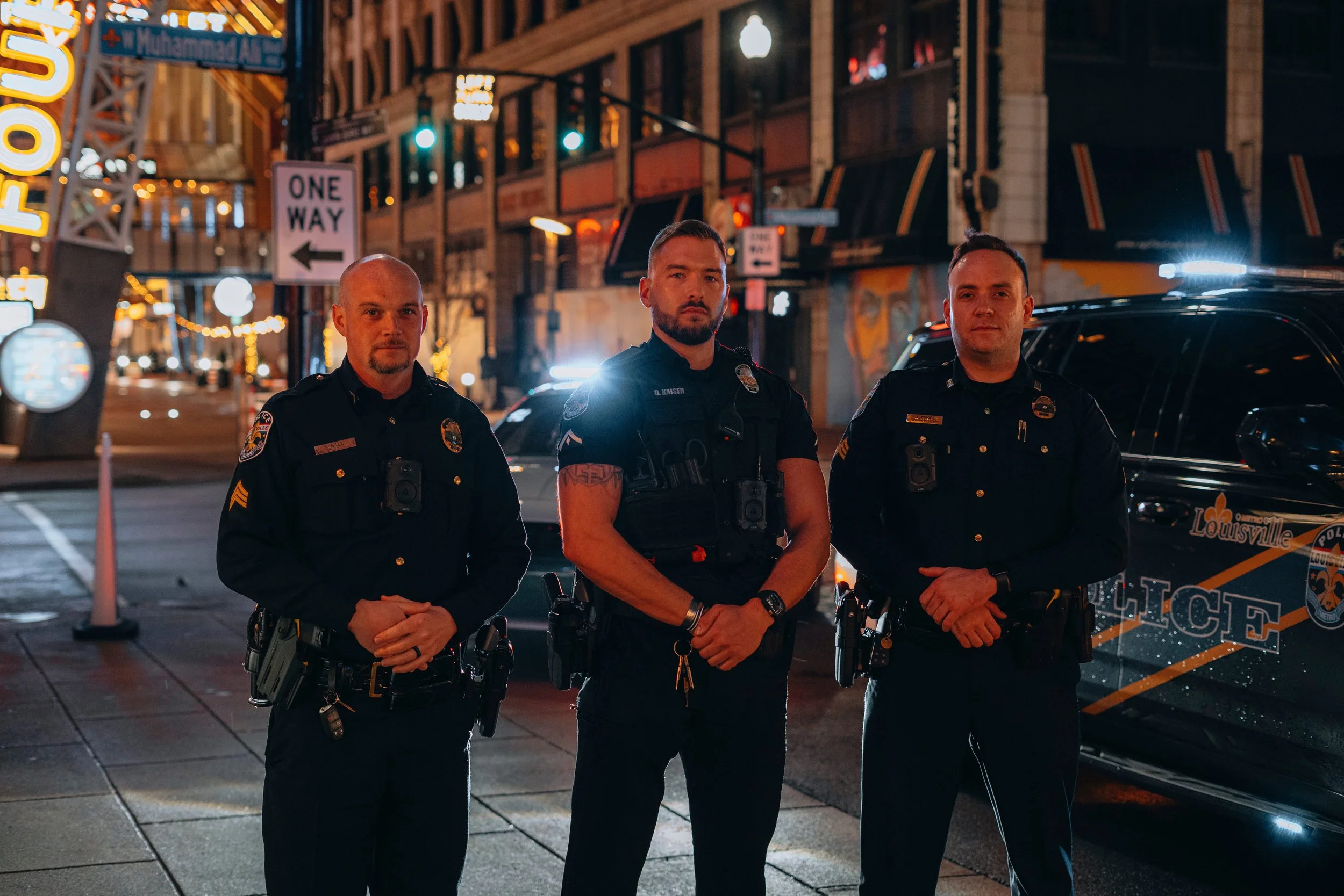 Three police officers standing on a city street at night, with buildings, streetlights, and police cars in the background.