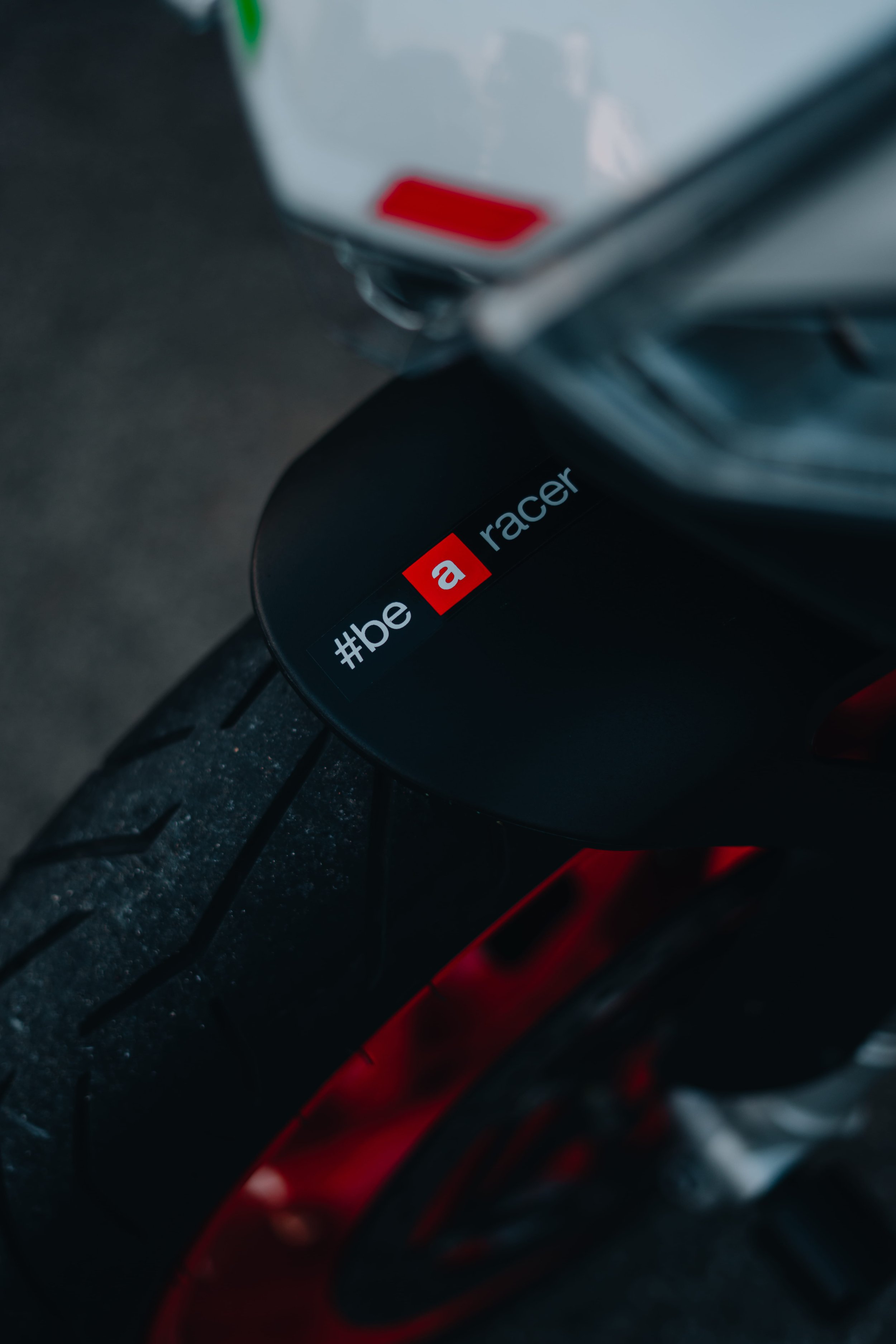Close-up of a black bicycle helmet with a red and white hashtag hashtag '#bikeracer' sticker on it, attached to a bicycle with a black tire and red accents, on a dark background.