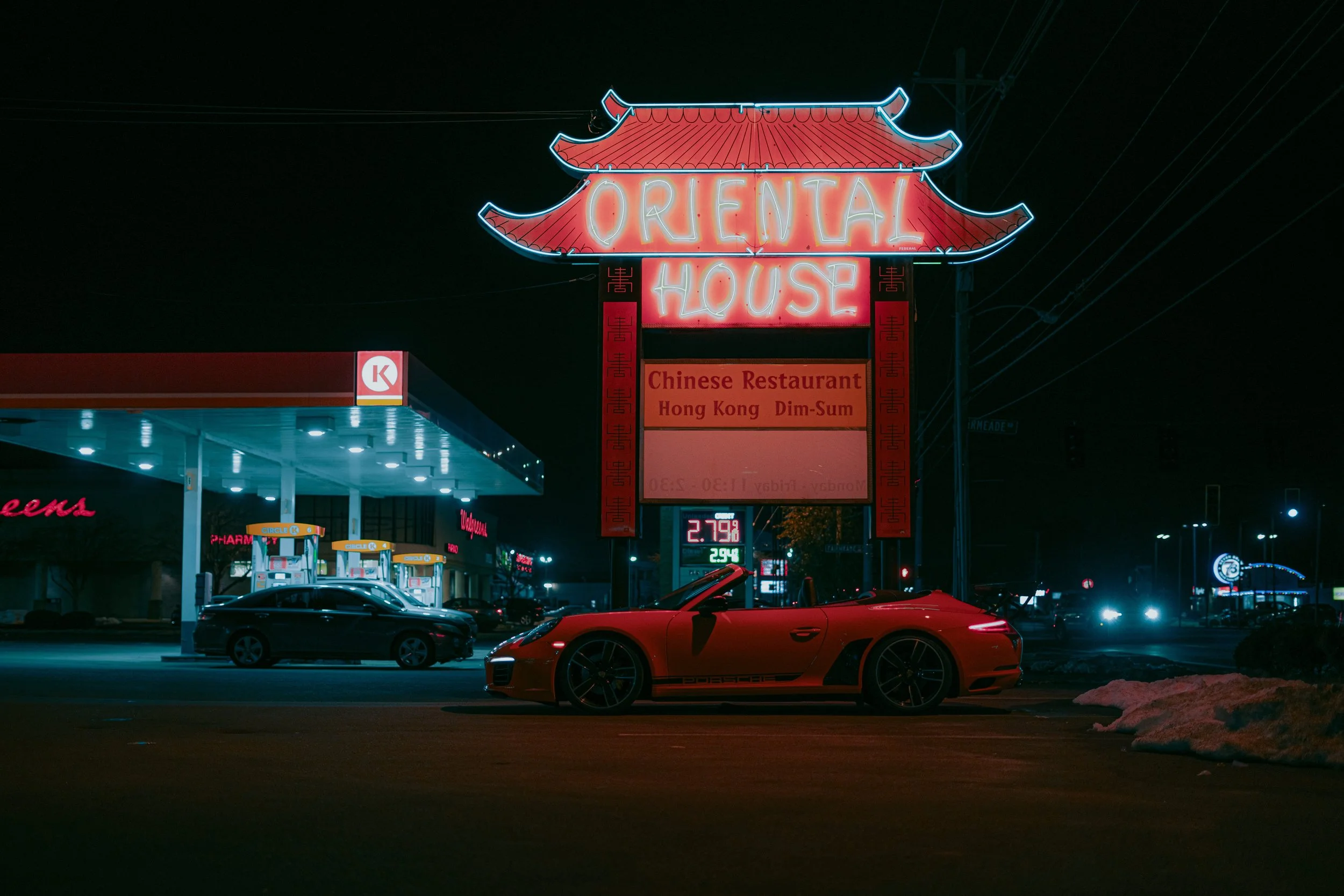 Nighttime scene of a neon sign for Orienta House Chinese Restaurant with Hong Kong Dim Sum. A red sports car is parked in front of the restaurant, and gas station pumps are visible nearby. There are some snow patches on the ground.