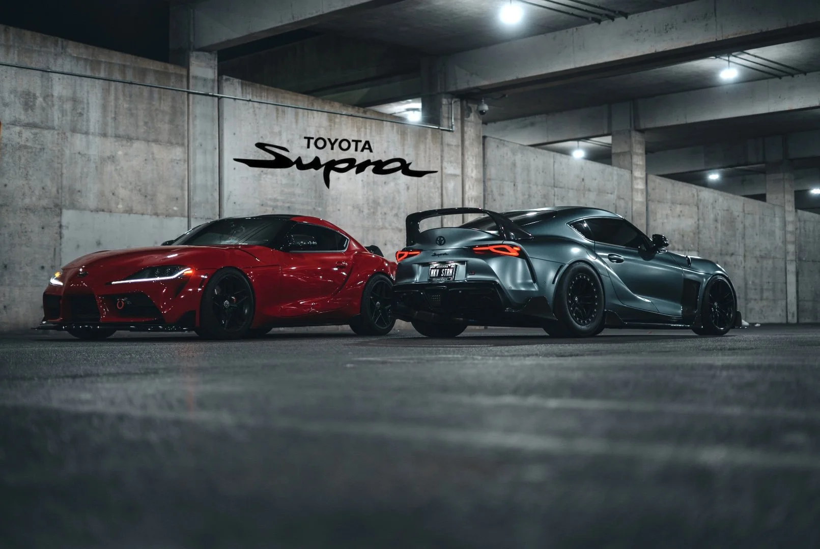 Two sports cars, one red Toyota Supra and one dark gray Toyota Supra, parked side by side in a dimly lit parking garage with a concrete wall featuring a "Toyota Supra" logo.