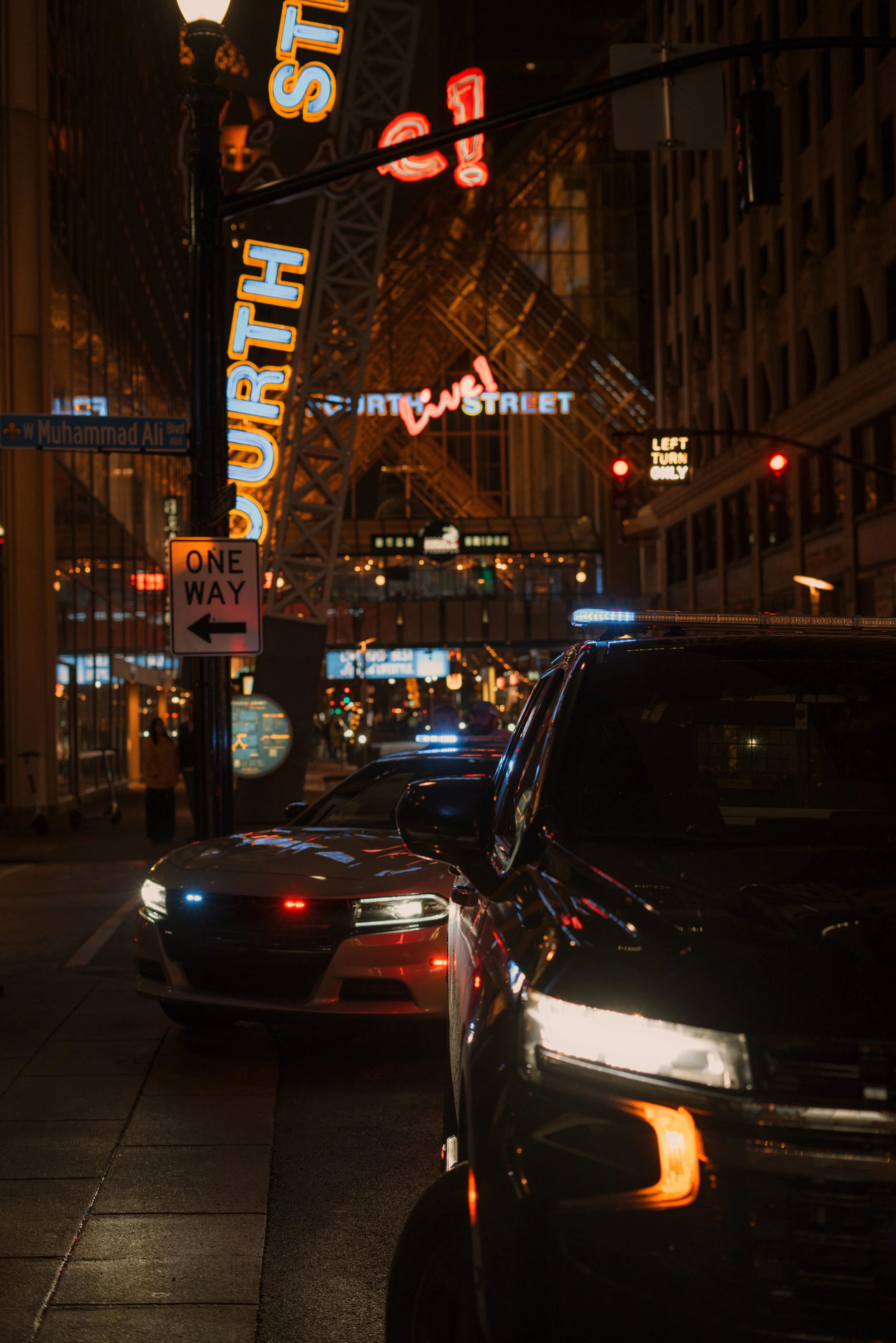 Nighttime city street scene with illuminated neon signs, including one that says "DURTY ST" and another with an exclamation mark. A police car and a dark sedan are parked on the street, with the police car's emergency lights on. A street sign reads "