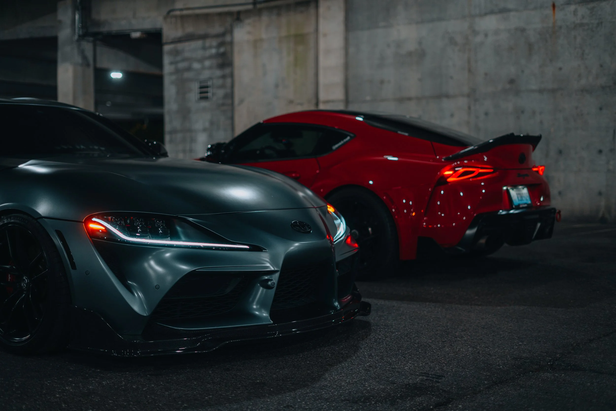 A silver sports car parked in front of a red sports car inside a concrete parking garage.