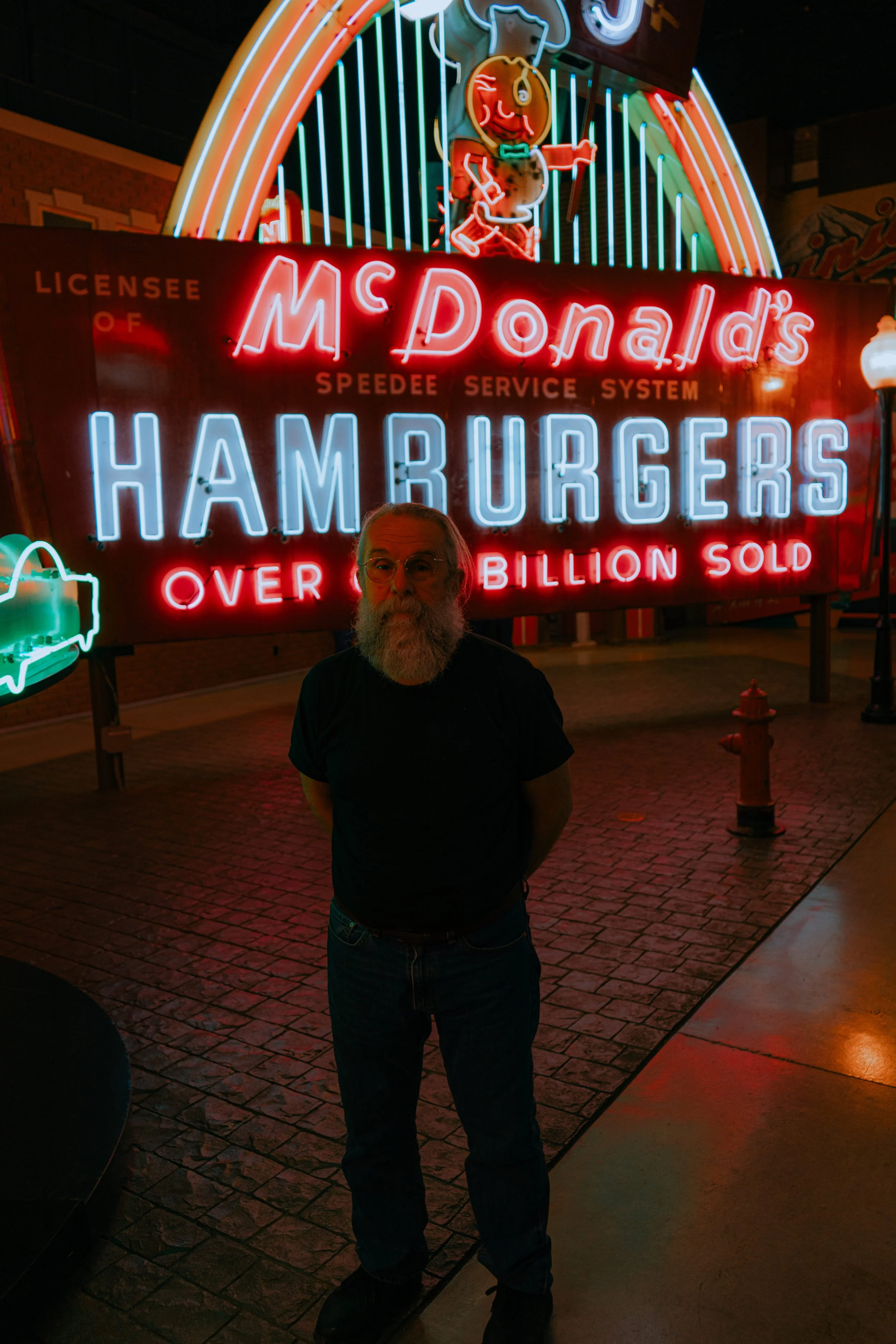 A man with glasses and a beard standing in front of a brightly lit neon sign for McDonald's hamburgers at night. The sign includes a cartoon character and colorful neon lights.