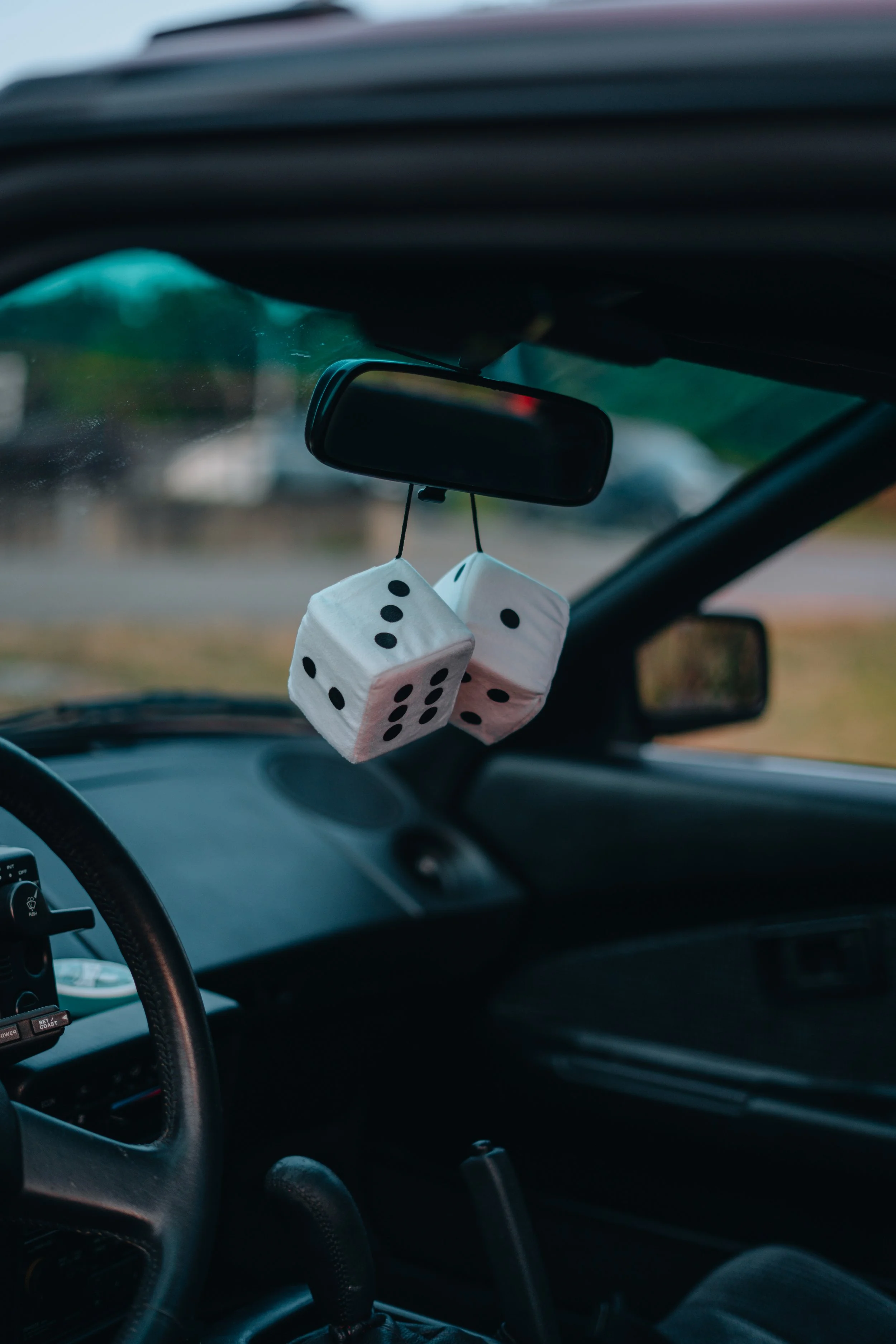 Inside a car showing a rearview mirror with two fuzzy white dice hanging from it, and part of the steering wheel and dashboard.