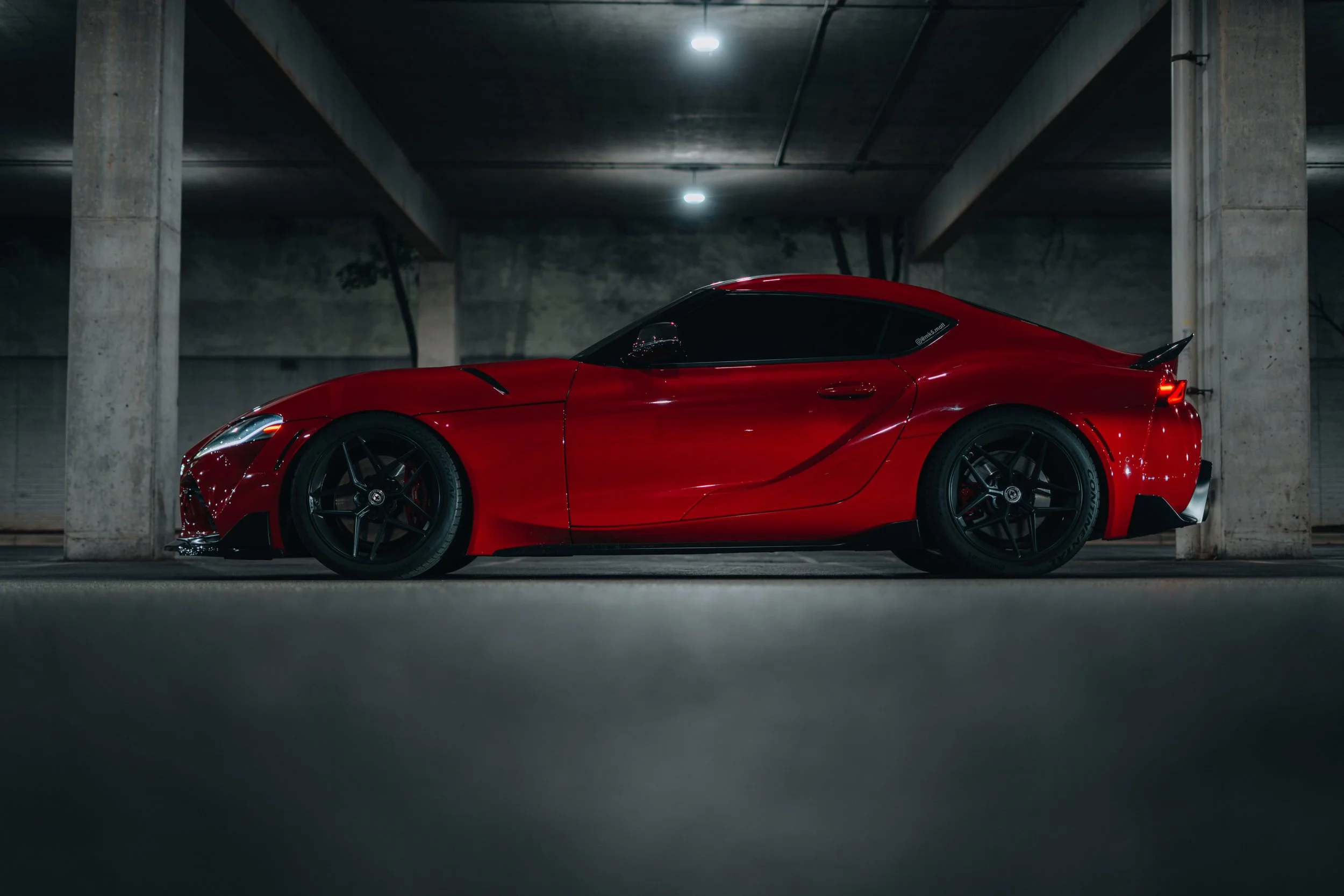 A red sports car parked in an underground parking garage with concrete pillars and a dark ceiling, illuminated by overhead lights.