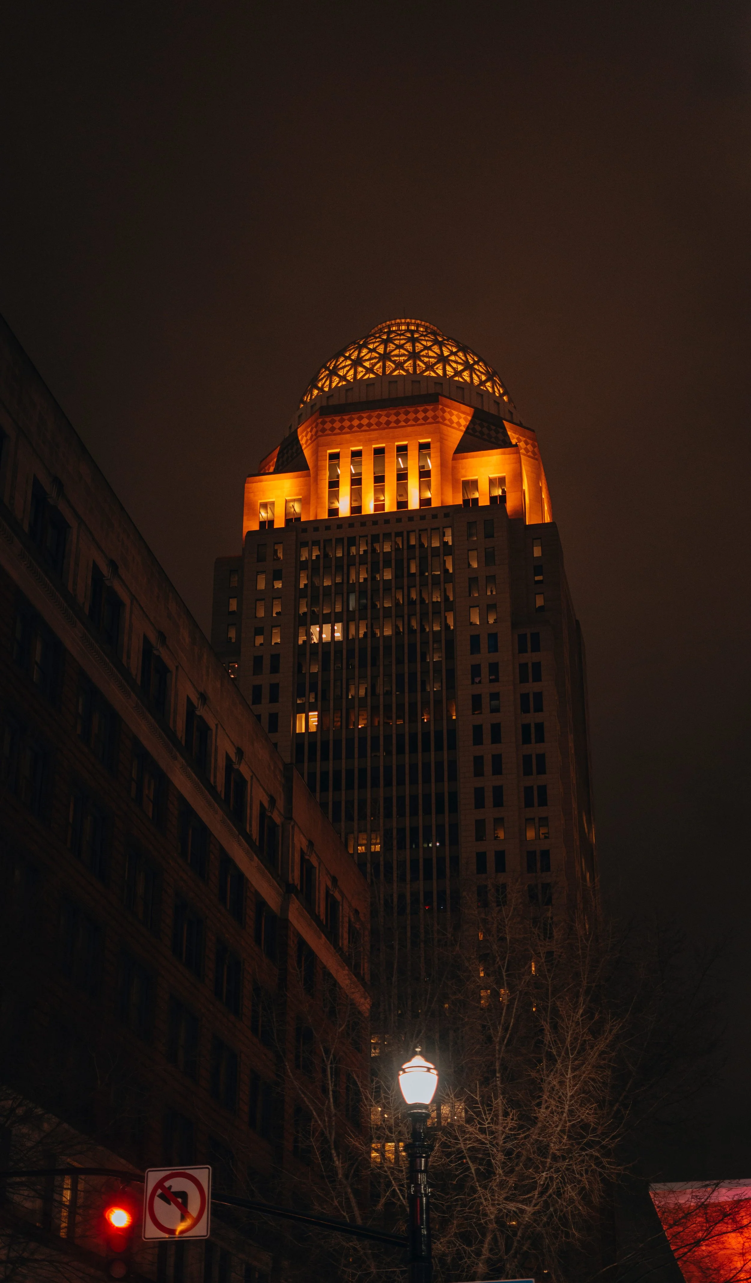 Nighttime city scene featuring a tall building illuminated with orange lights and a glass-domed top, a streetlamp, and a no U-turn traffic sign.